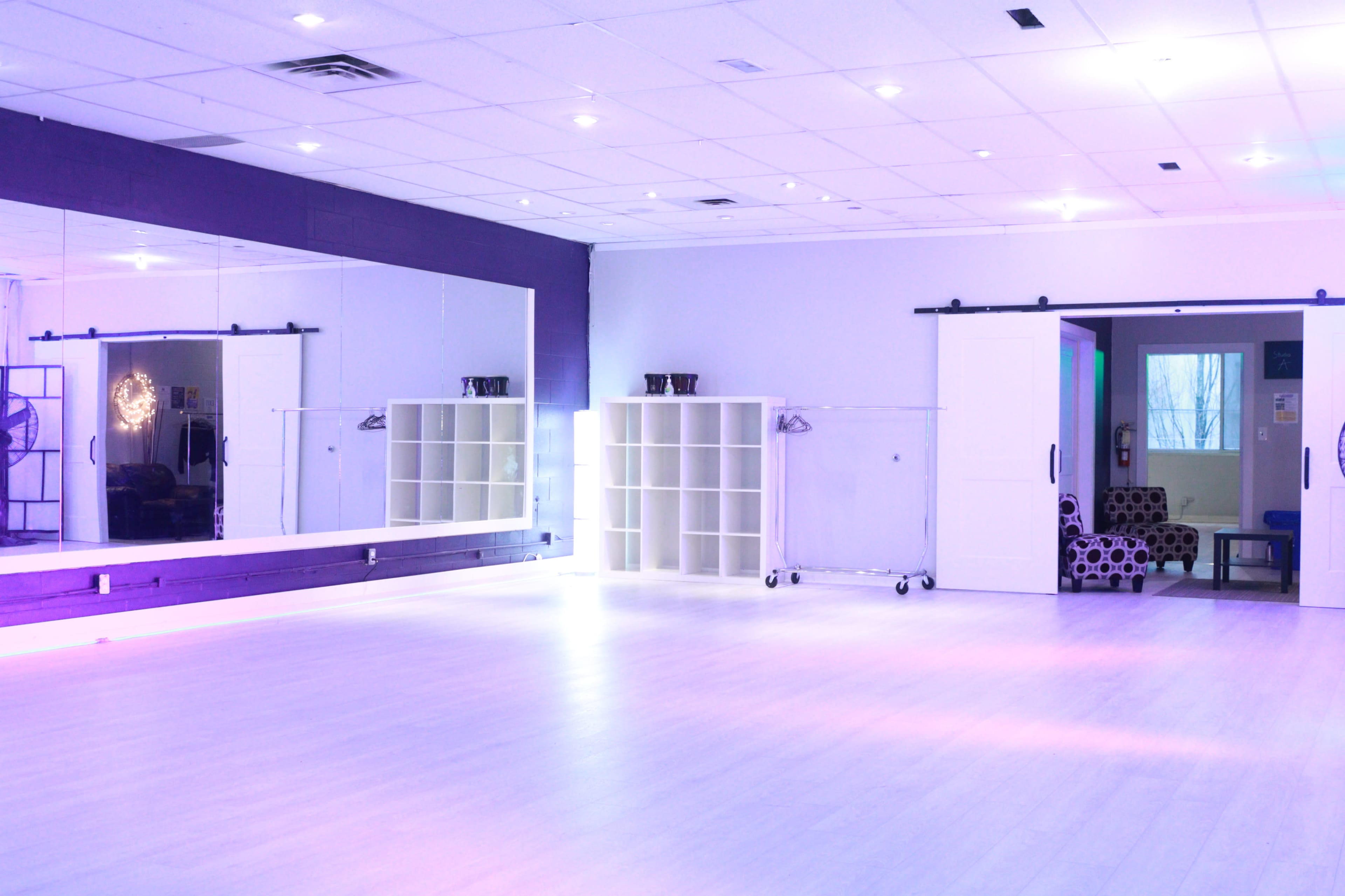 A brightly lit dance studio features a large mirrored wall, shelving unit, and a cozy seating area in the background.