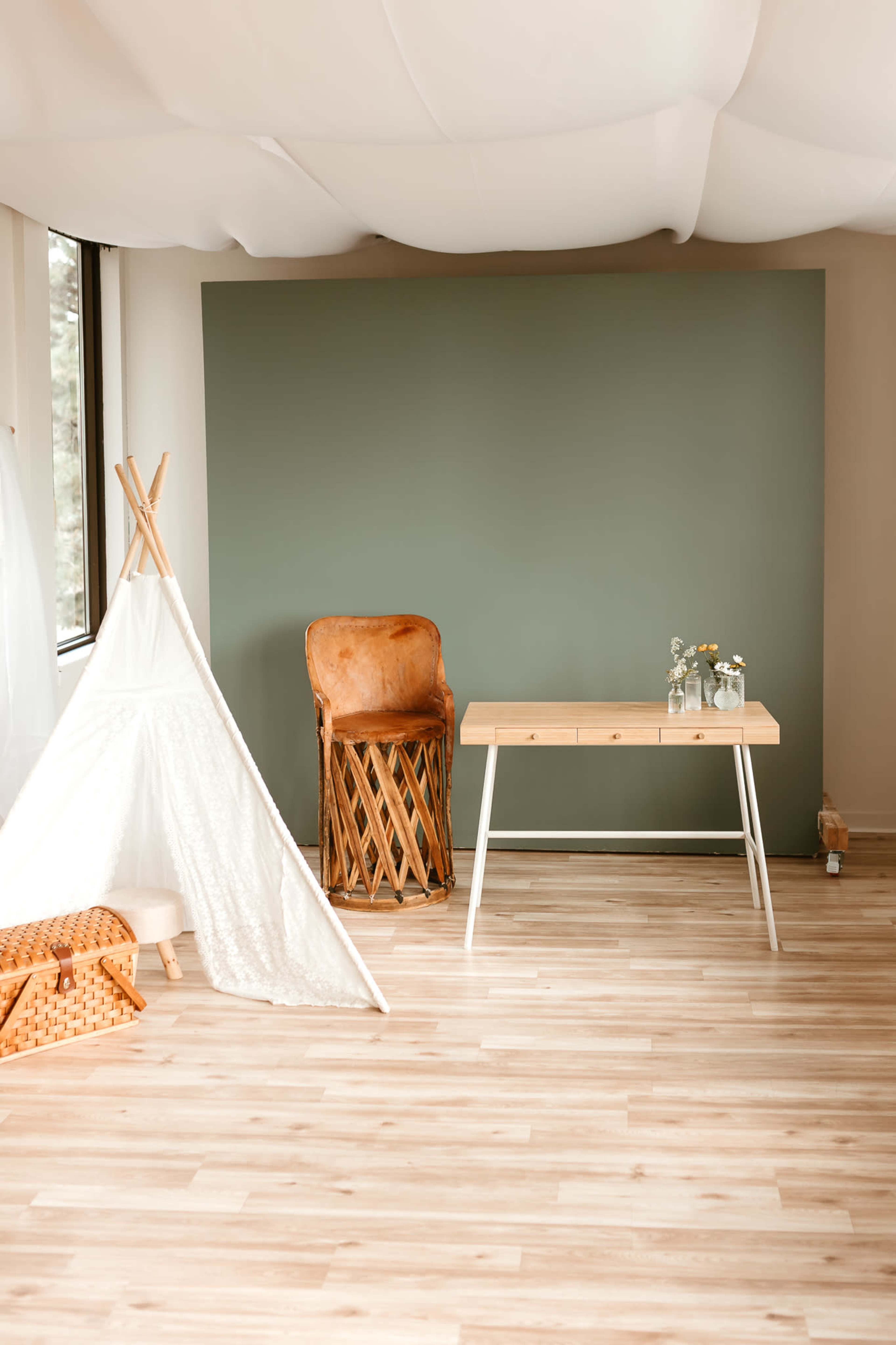 A minimalist room with a green wall, a wooden table, a leather chair, a white teepee, and a wicker basket on the floor.