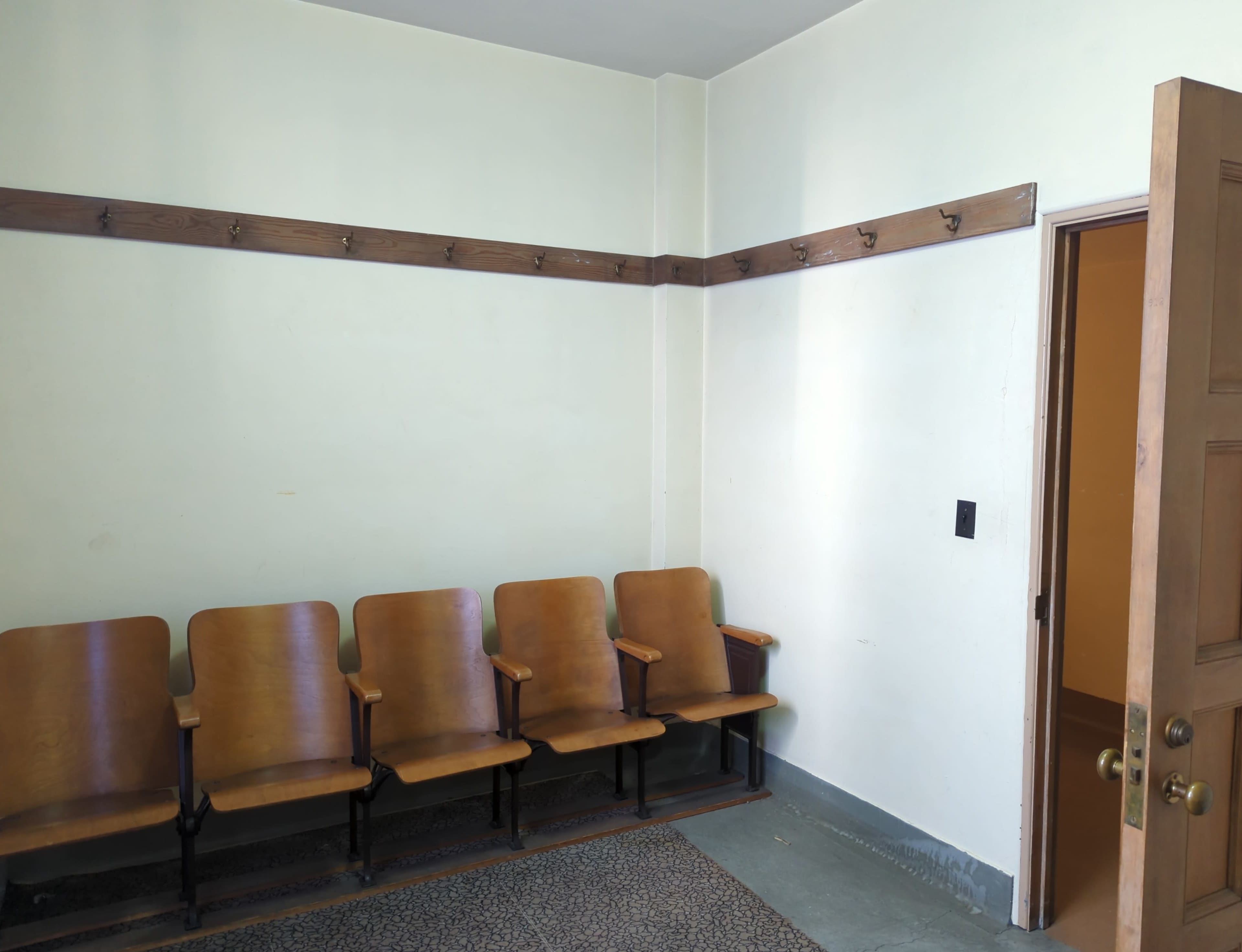 The image shows a small room with a row of four brown chairs against a pale wall, a coat rack mounted above them, and a door on the right.