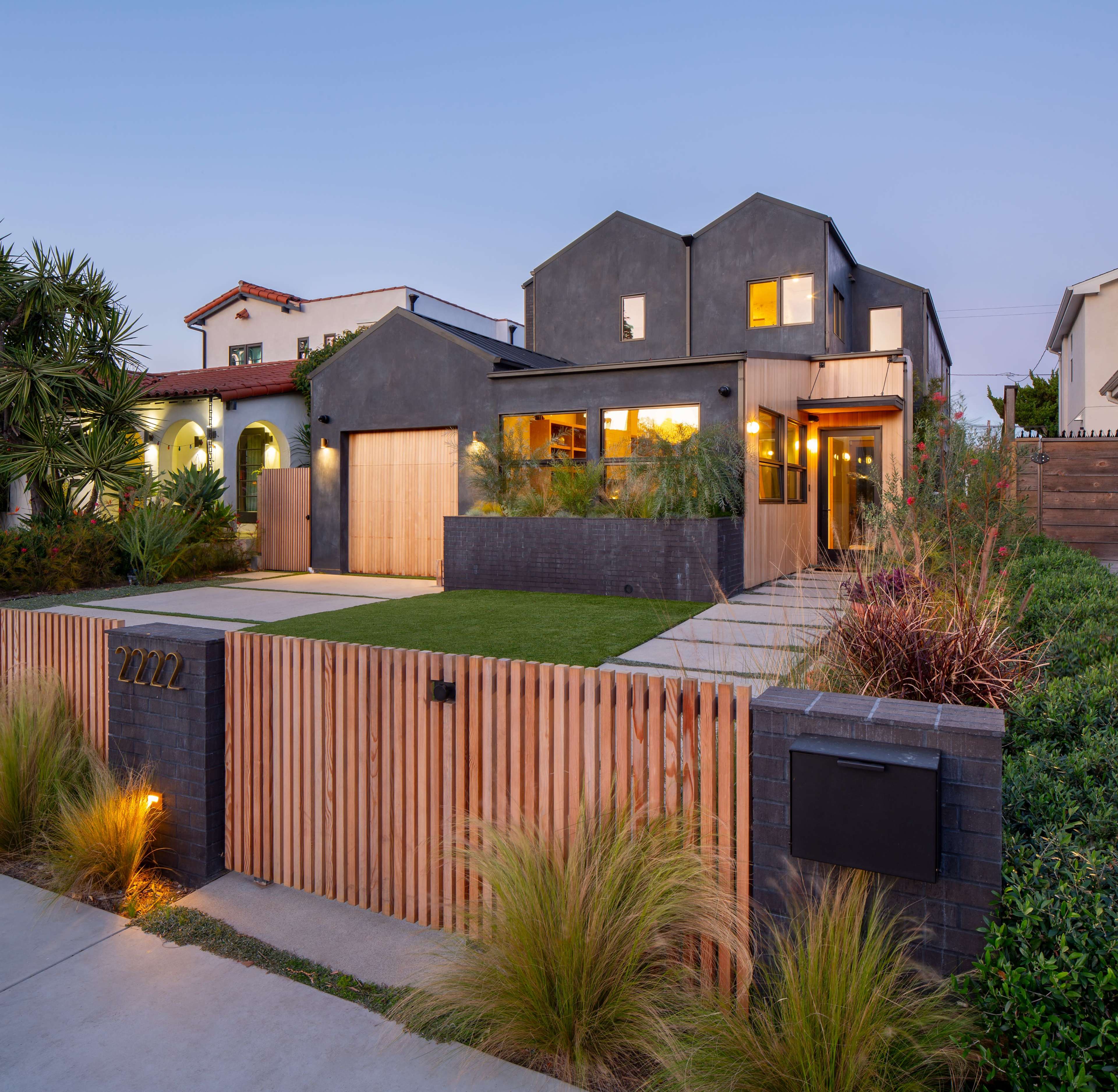 A modern house with a wooden fence and landscaped front yard is shown at dusk, featuring large windows and a combination of gray and wooden exterior accents.