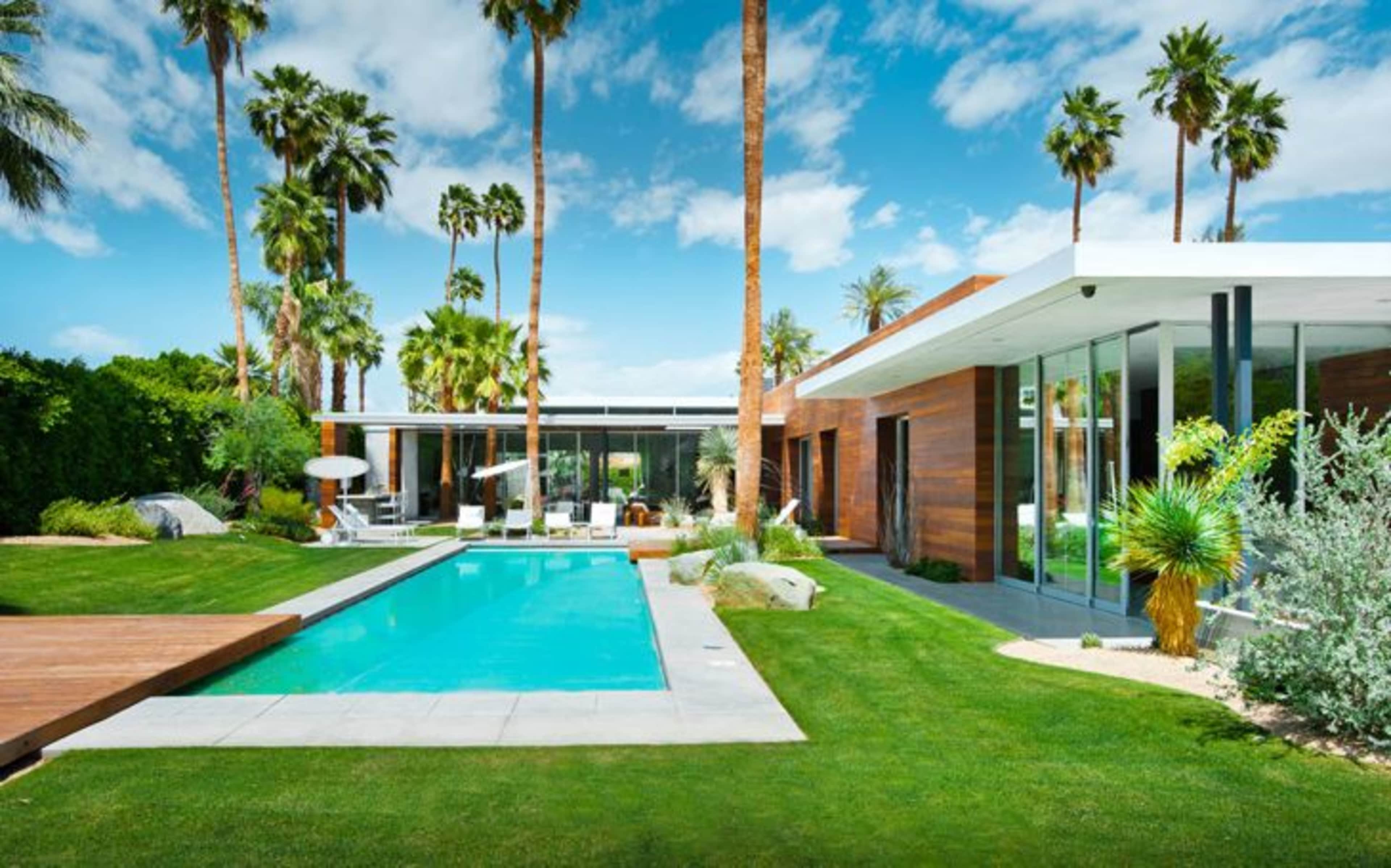 A modern home with large windows and a swimming pool is surrounded by palm trees and well-manicured grass.
