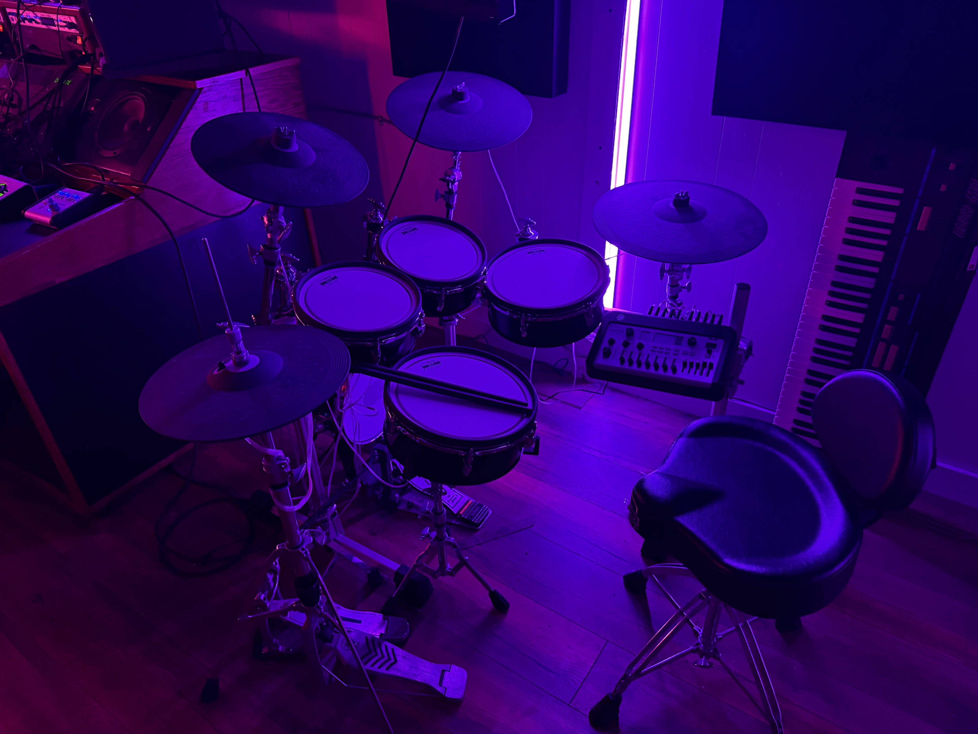 The image shows an electronic drum set with a stool in a room illuminated by purple lighting.