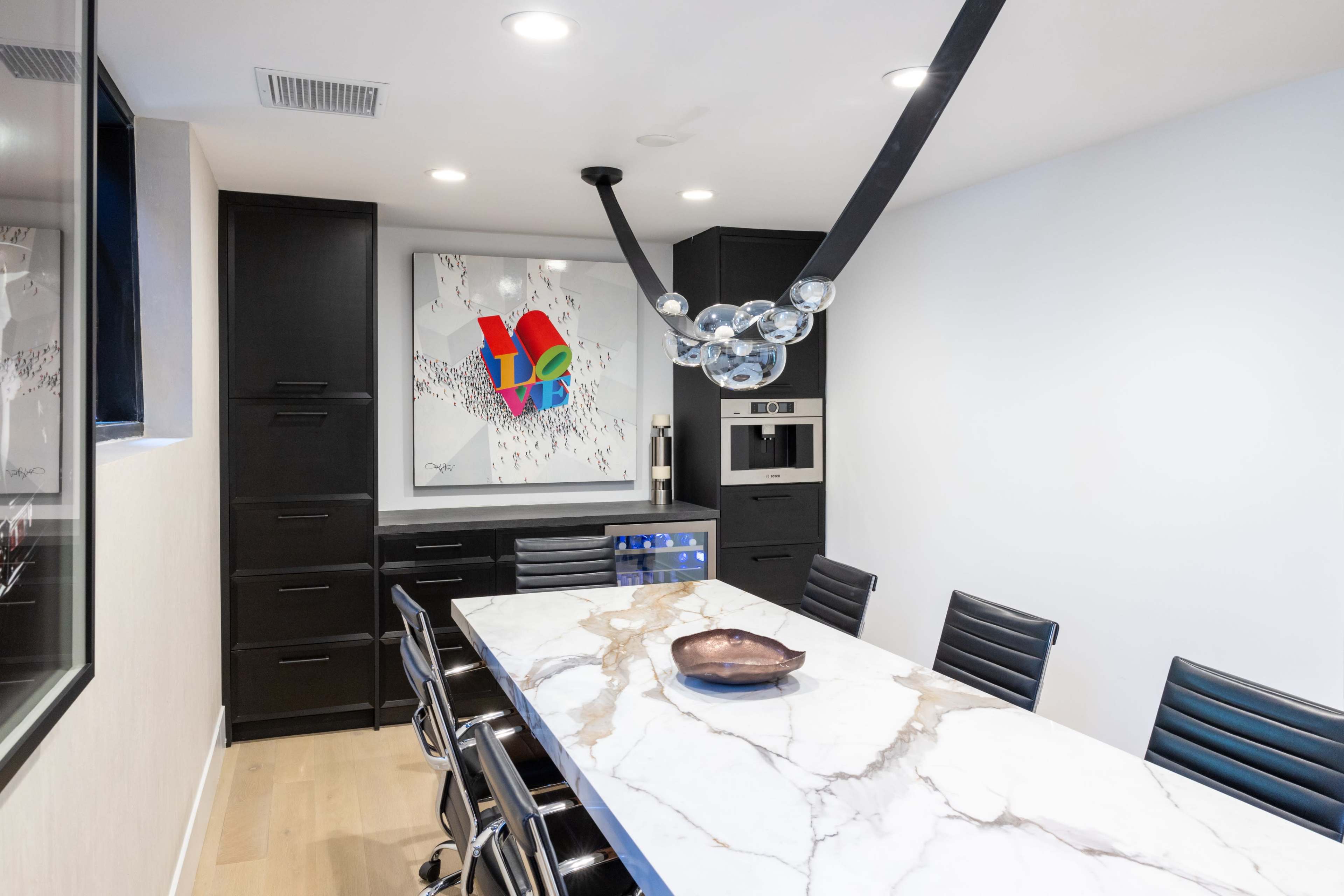 A modern dining area features a long marble table surrounded by black chairs, with a colorful art piece and a stylish light fixture overhead.