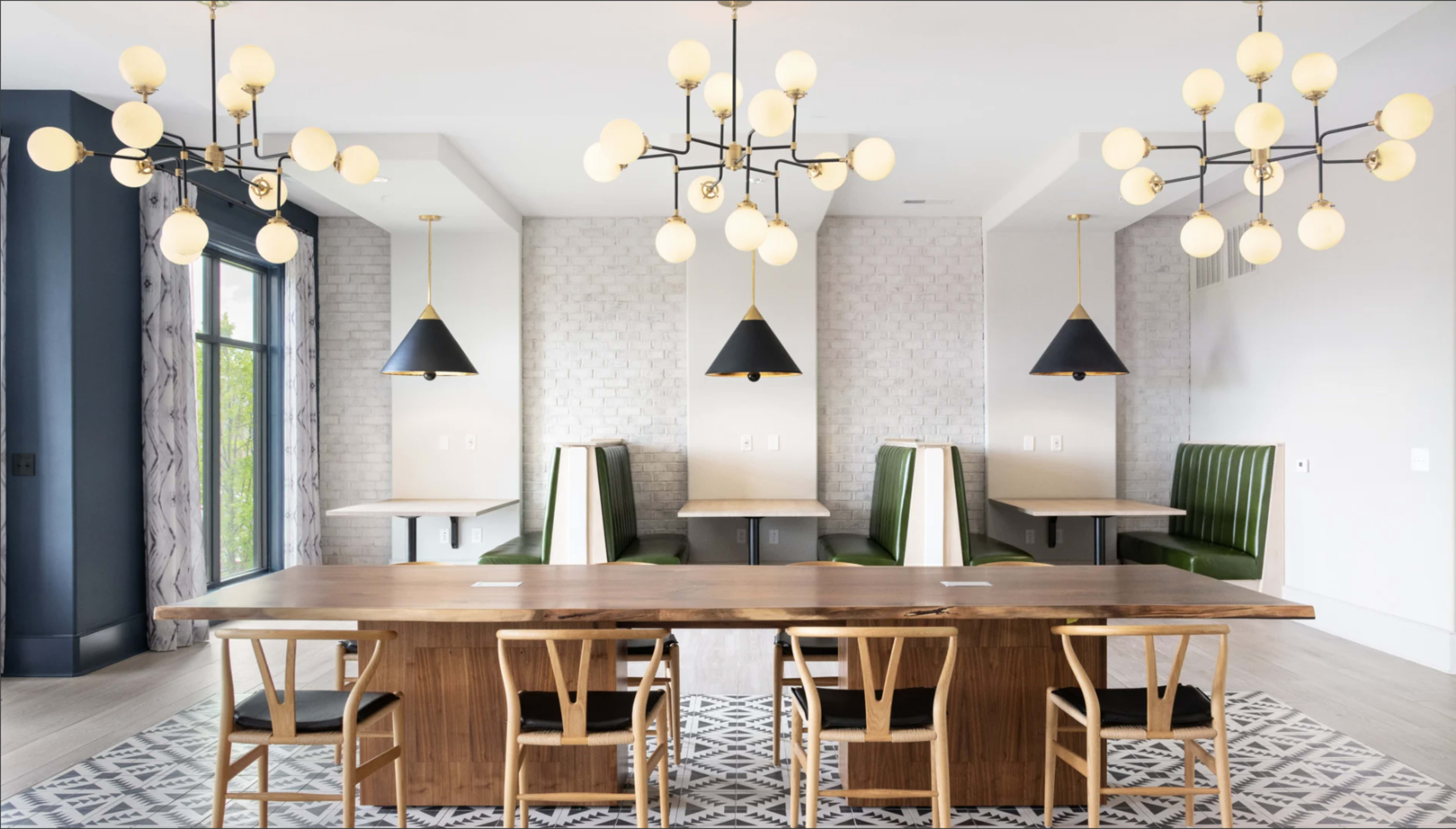 The image shows a modern dining area with a long wooden table, several black and gold light fixtures, and green upholstered booths along the walls.