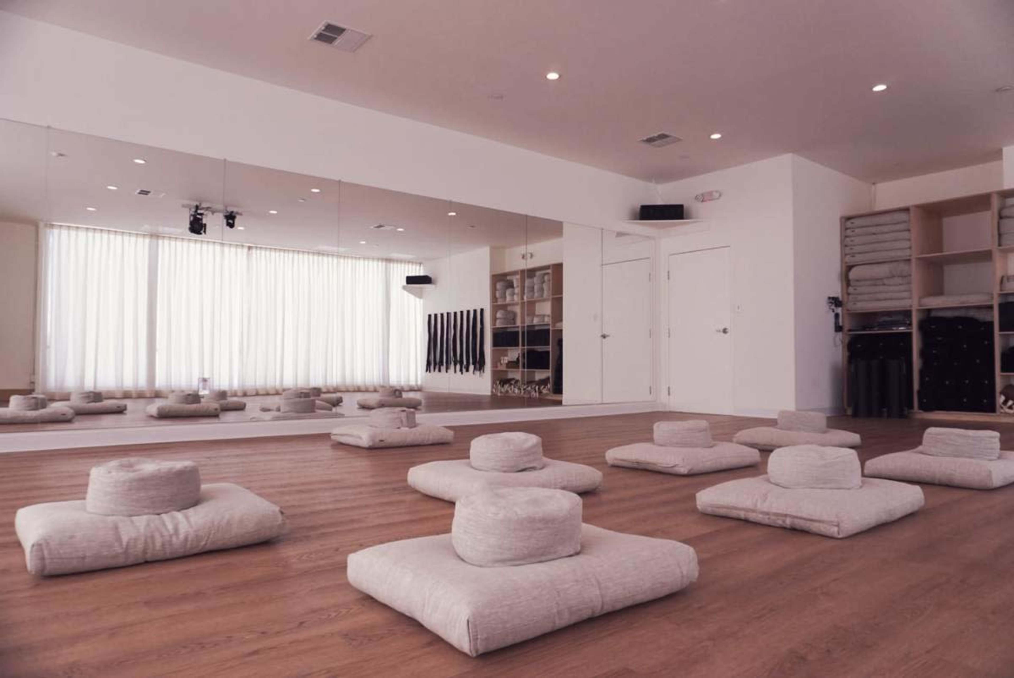 The image shows a quiet yoga or meditation studio with several floor cushions arranged on a wooden floor, and large windows allowing natural light to enter.