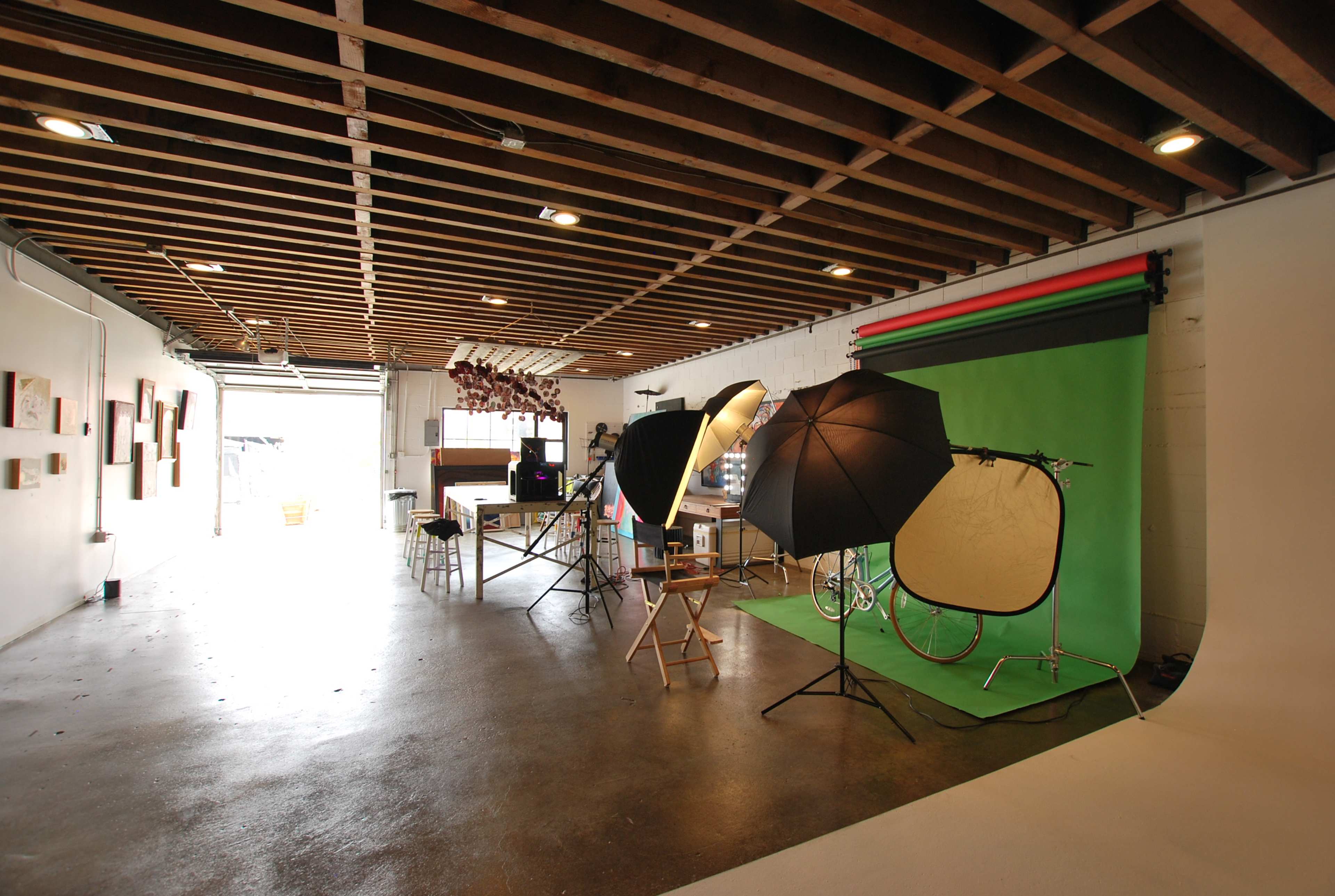 The image shows a photo studio with lighting equipment, a green backdrop, and various props arranged in a spacious room with exposed wooden beams.