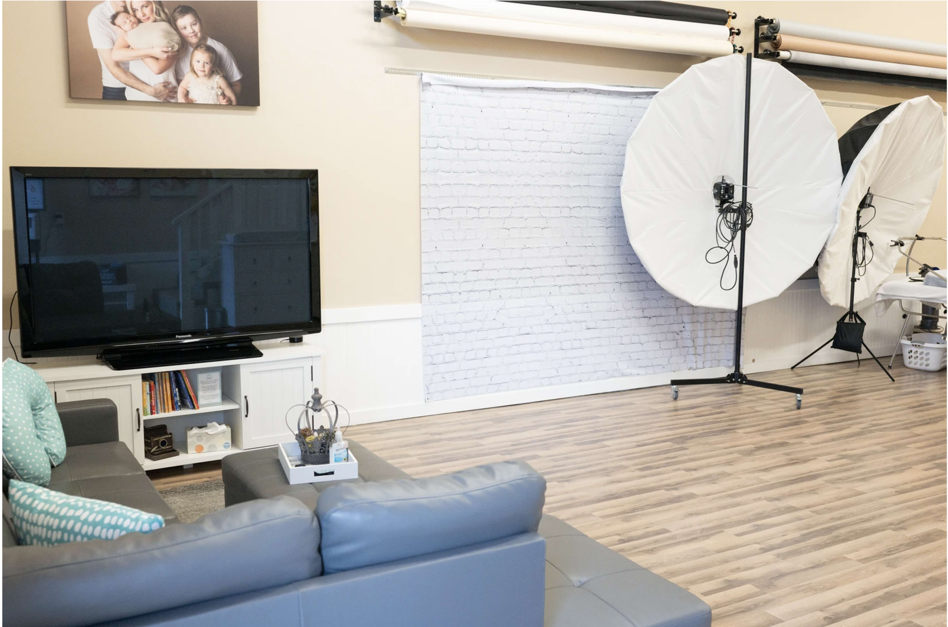 The image shows a bright interior space featuring a gray sofa, a television, and large photography lights set up next to a brick-patterned backdrop.