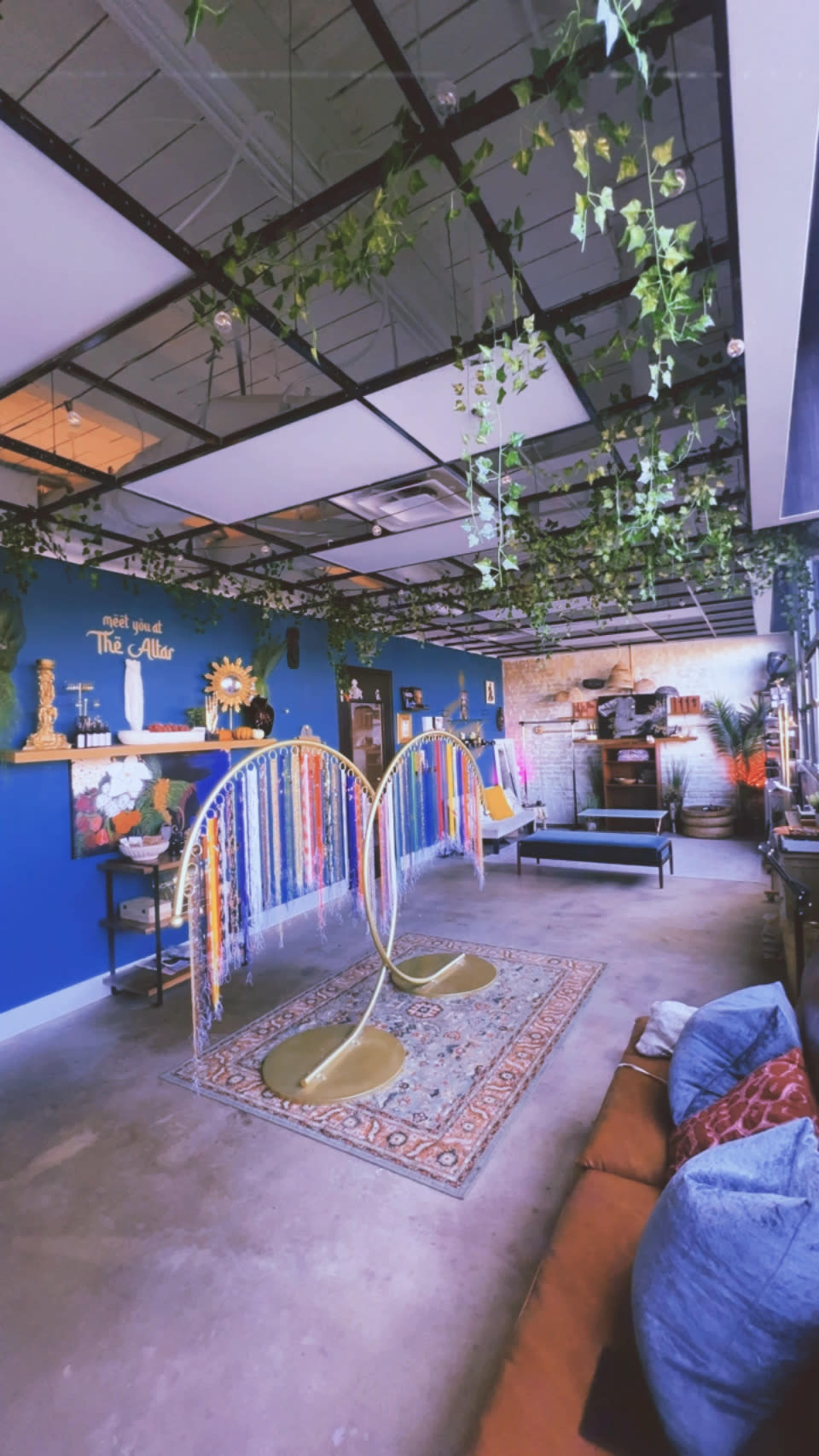 The image shows a vibrant indoor space featuring colorful hanging decor, plants adorning the ceiling, and several seating areas arranged around a patterned rug.