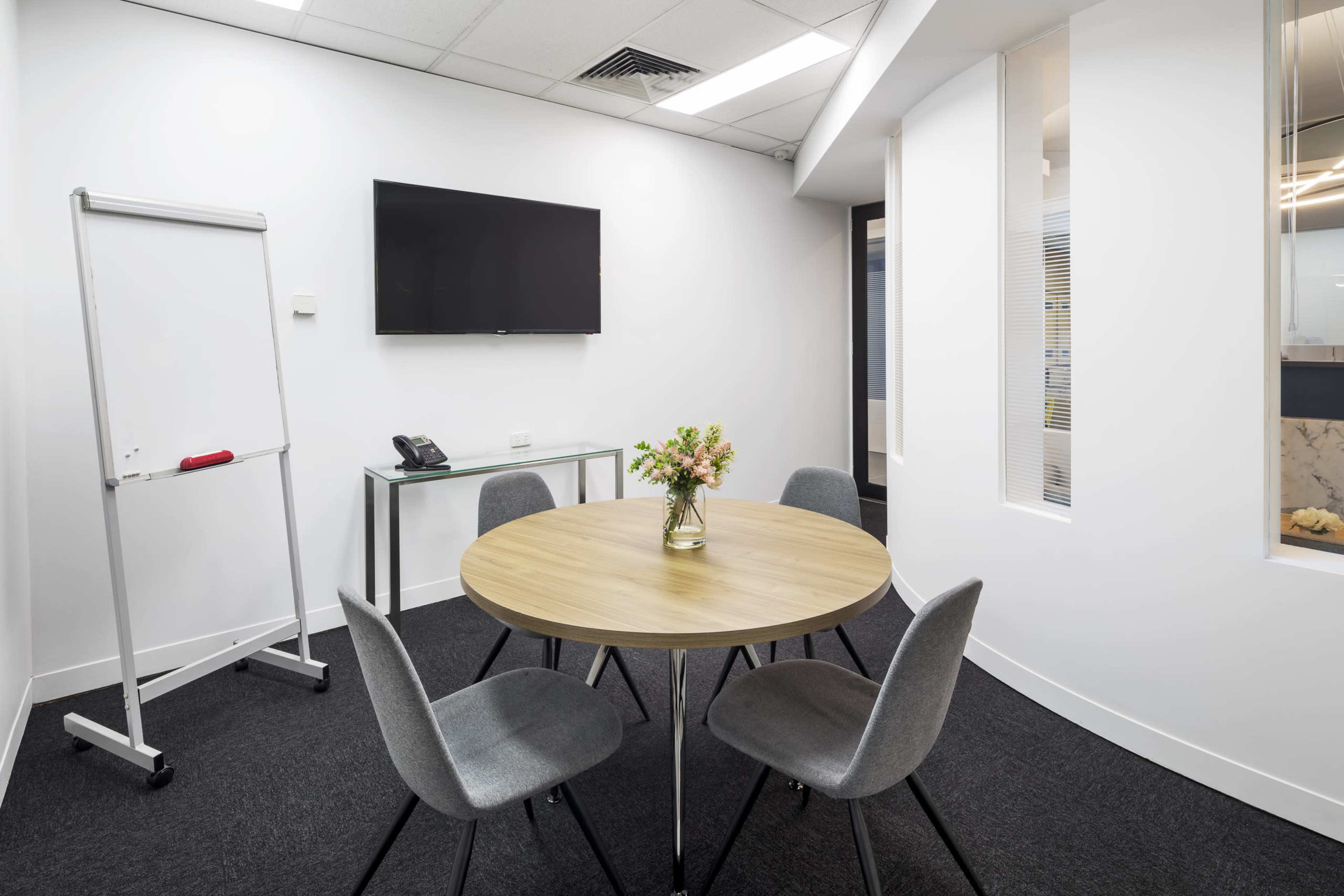 A small meeting room features a round wooden table surrounded by four gray chairs, a television mounted on the wall, a whiteboard, and a vase of flowers on the table.
