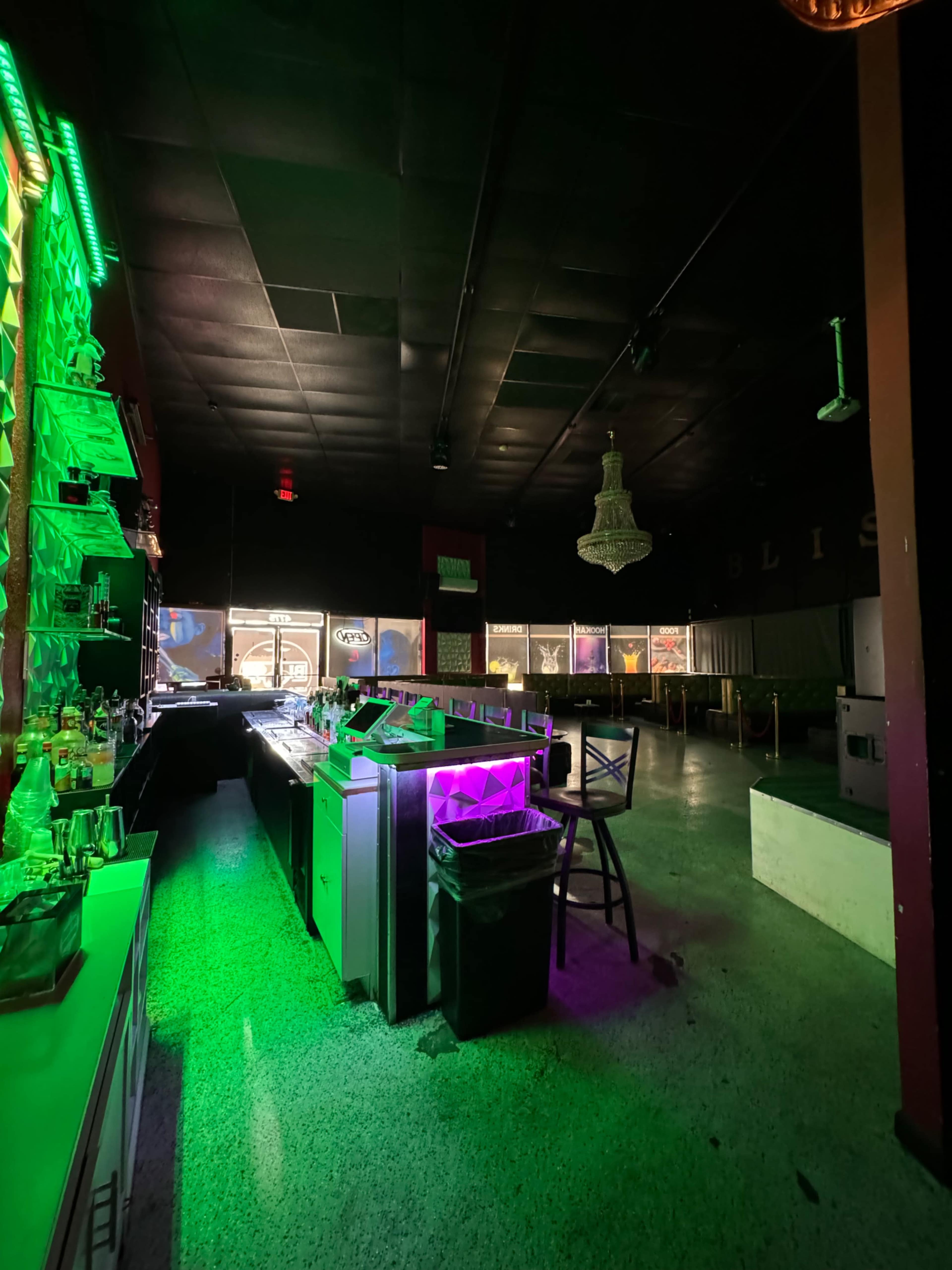 An empty bar area with green and purple lighting, featuring a counter and a chandelier in a dimly lit space.