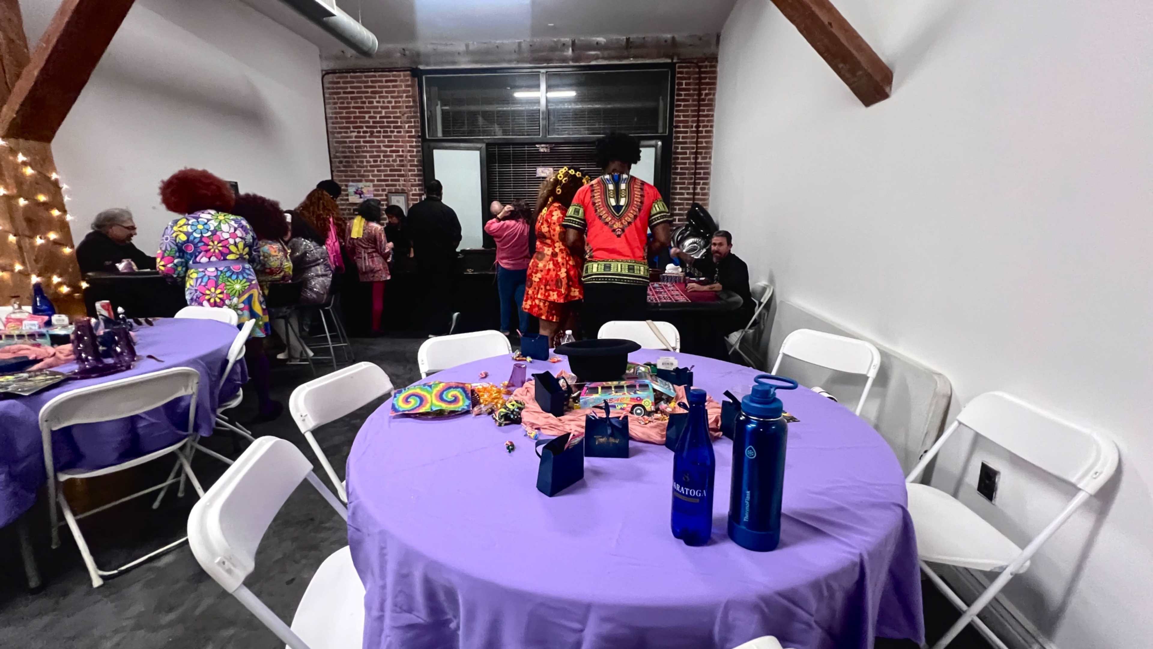 The image shows a gathering in a room with purple tablecloths on round tables, where individuals dressed in colorful attire are engaged in conversation.