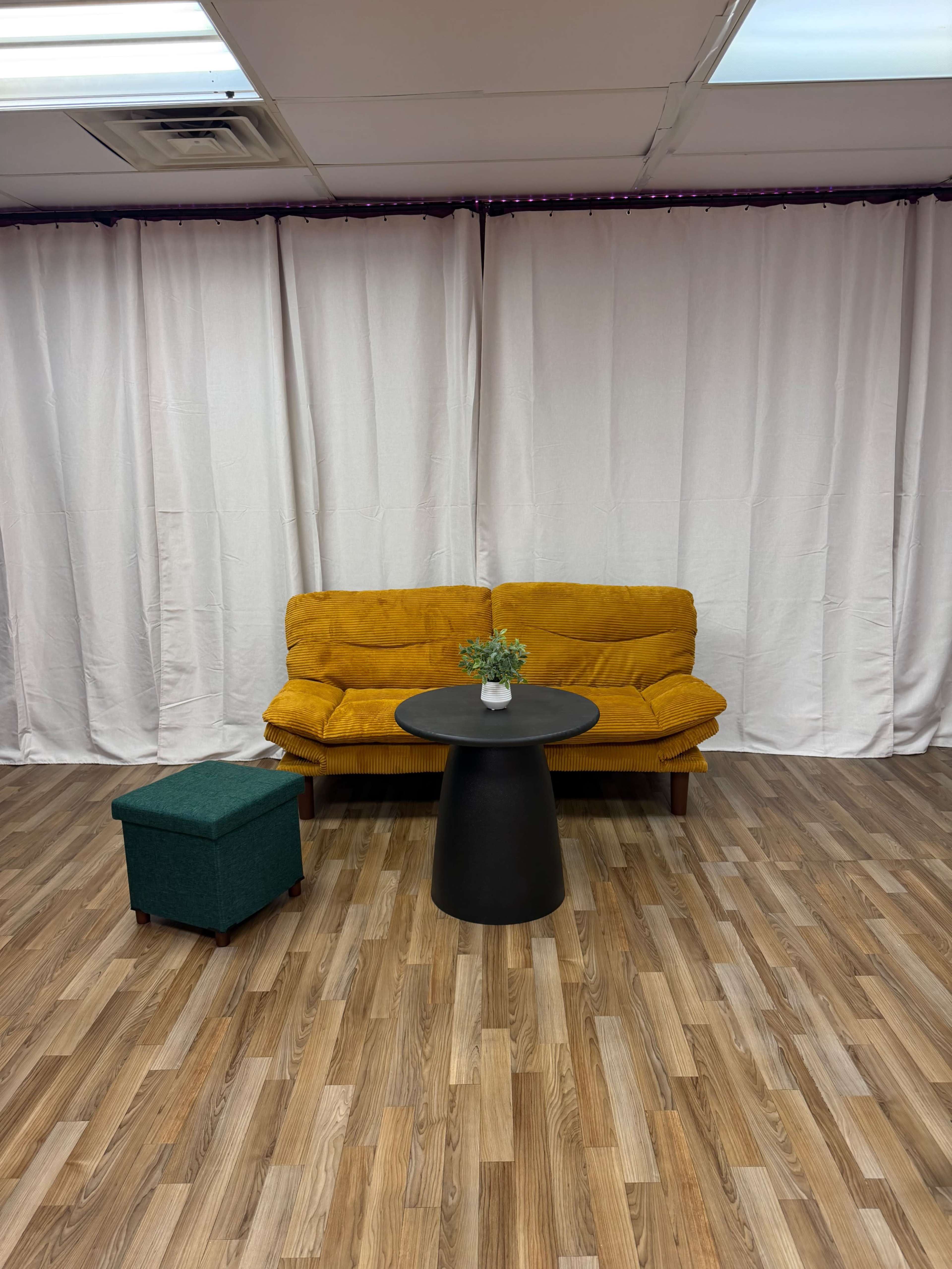 The image shows a yellow couch facing a round black table in a room with white curtains and a wooden floor.