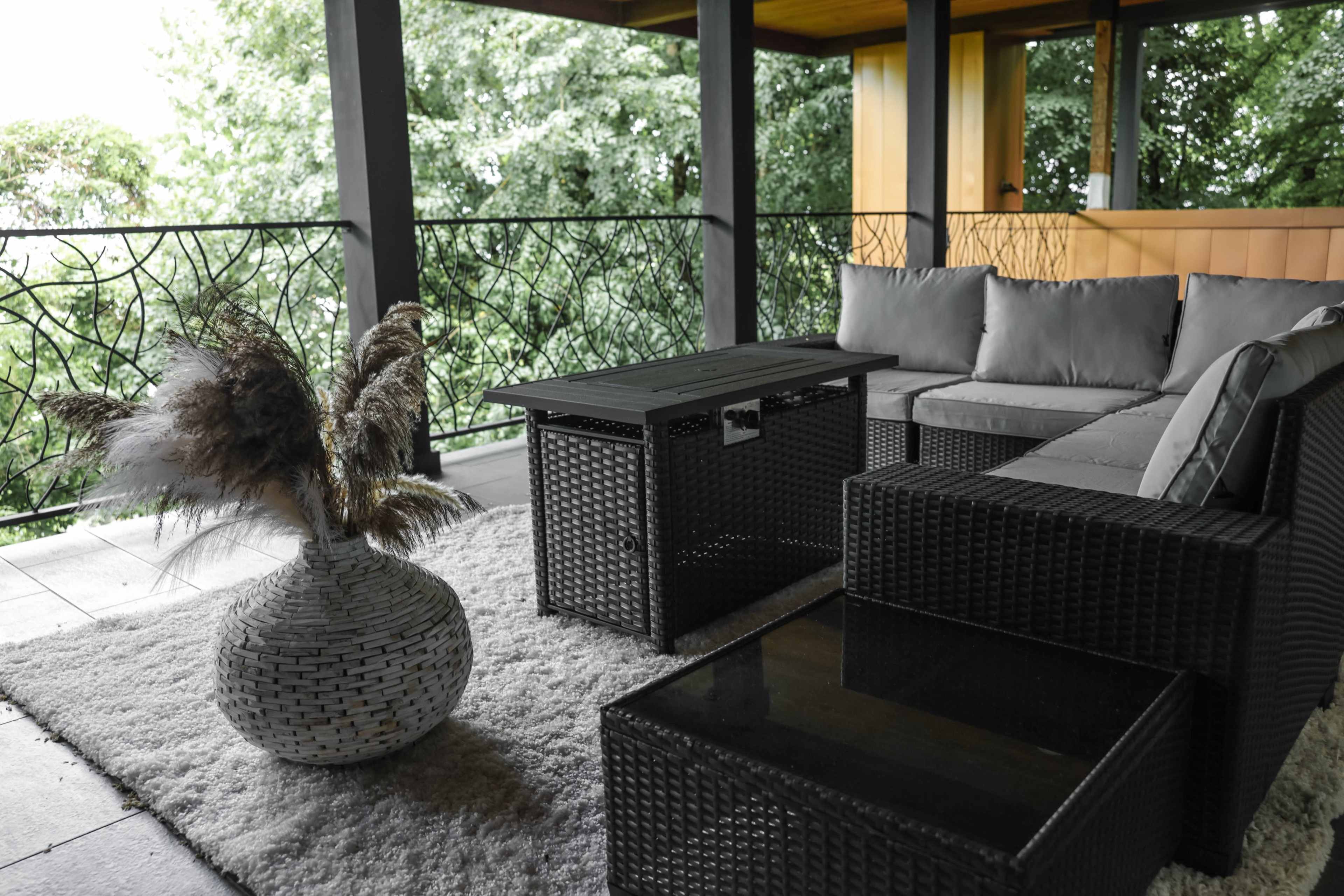 The image shows a modern outdoor seating area with wicker furniture, a small table, and a decorative vase in front of a view of greenery.
