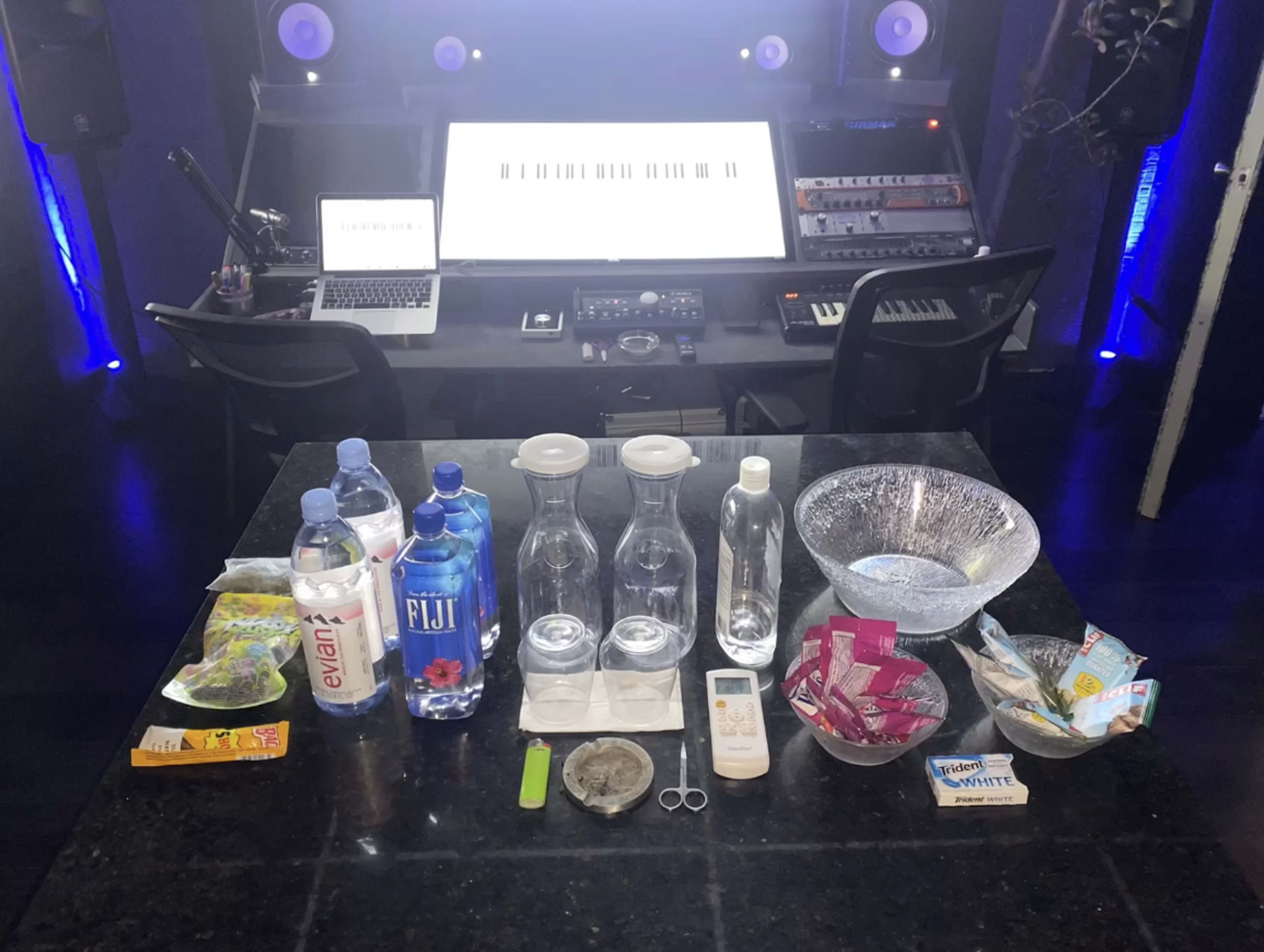 A recording studio setup with a large screen, musical equipment, and a table displaying various bottles of water, a bowl, a thermometer, scissors, and snacks.