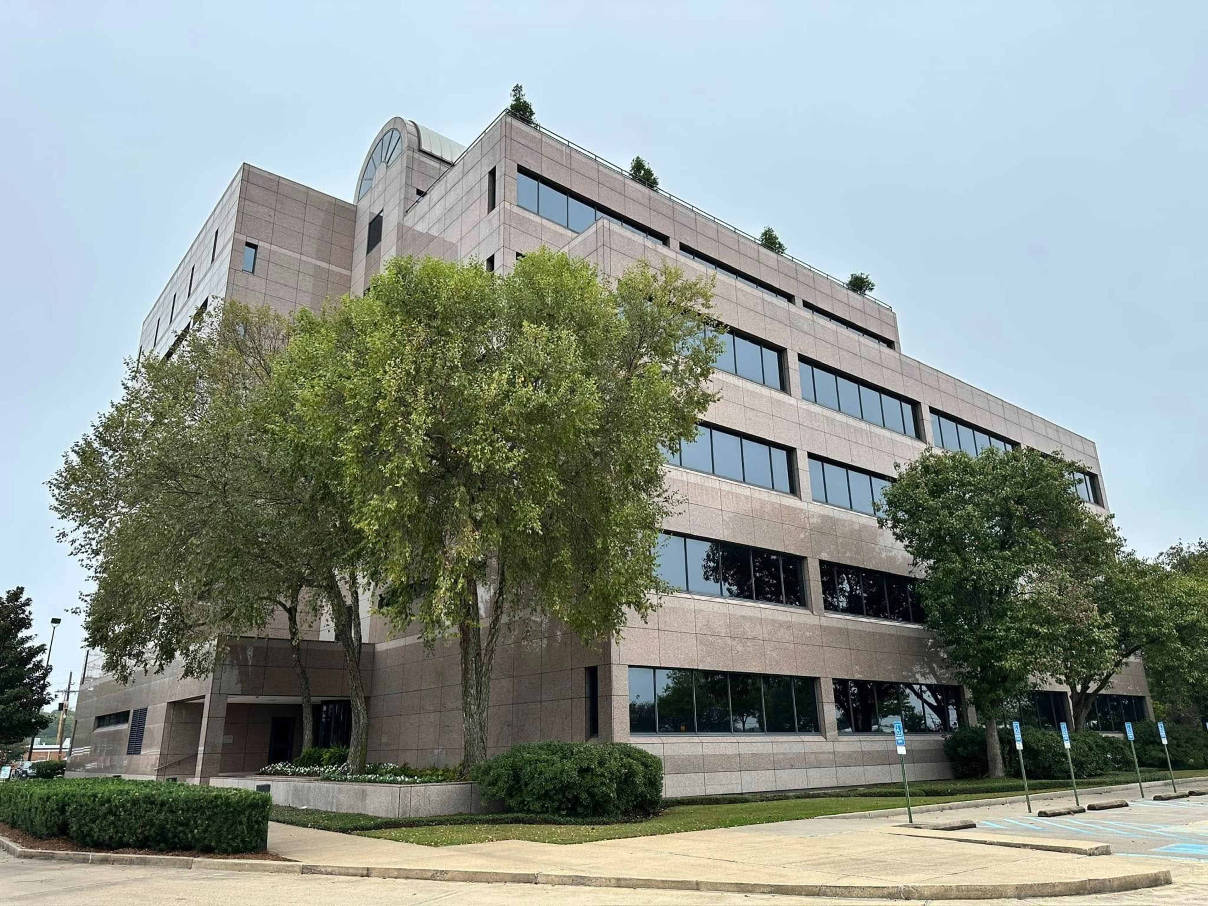 A multi-story office building with large windows and greenery at its base is set against a cloudy sky.