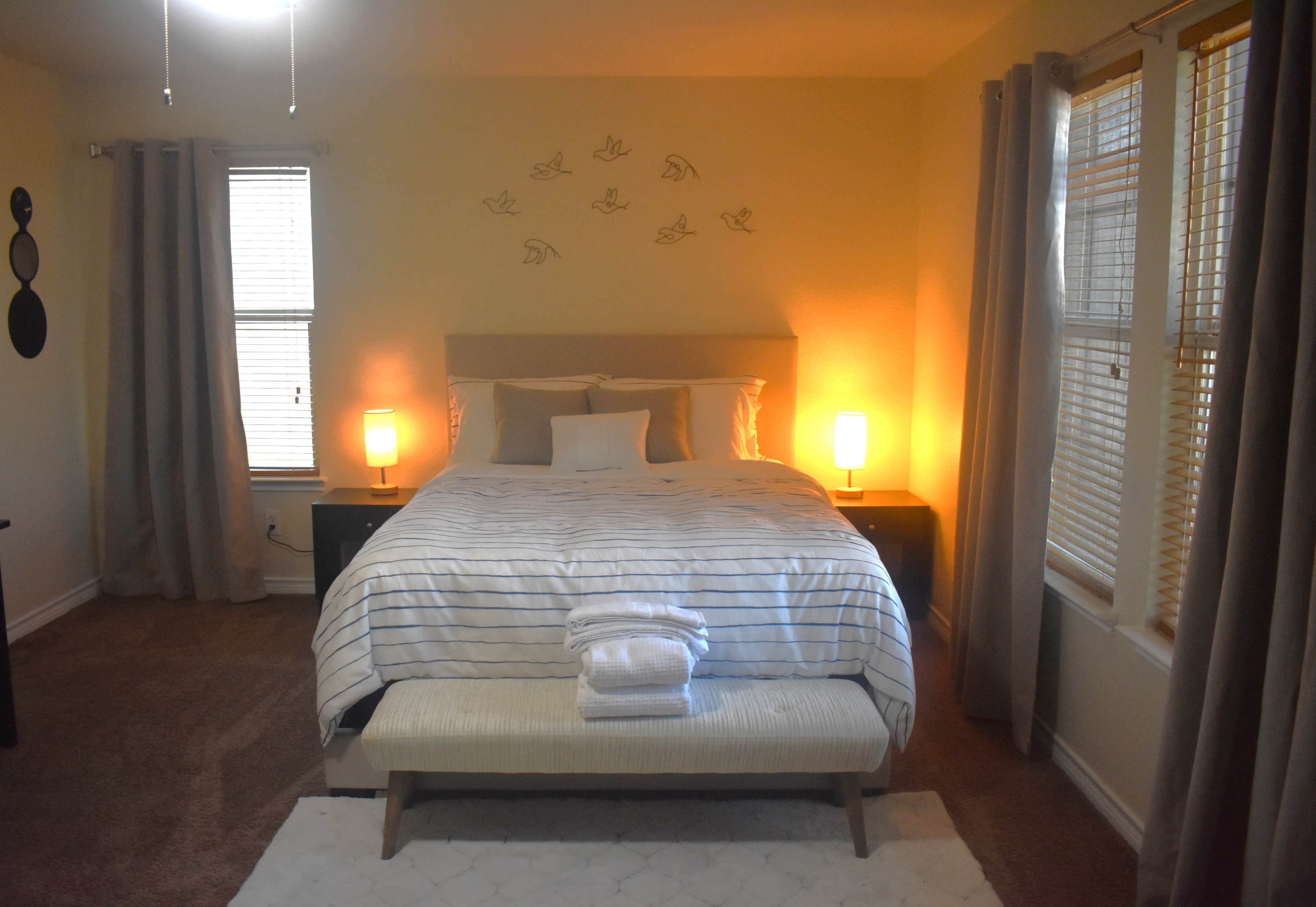 A neatly arranged bedroom features a bed with white and striped bedding, two lamps on nightstands, and a bench at the foot of the bed.