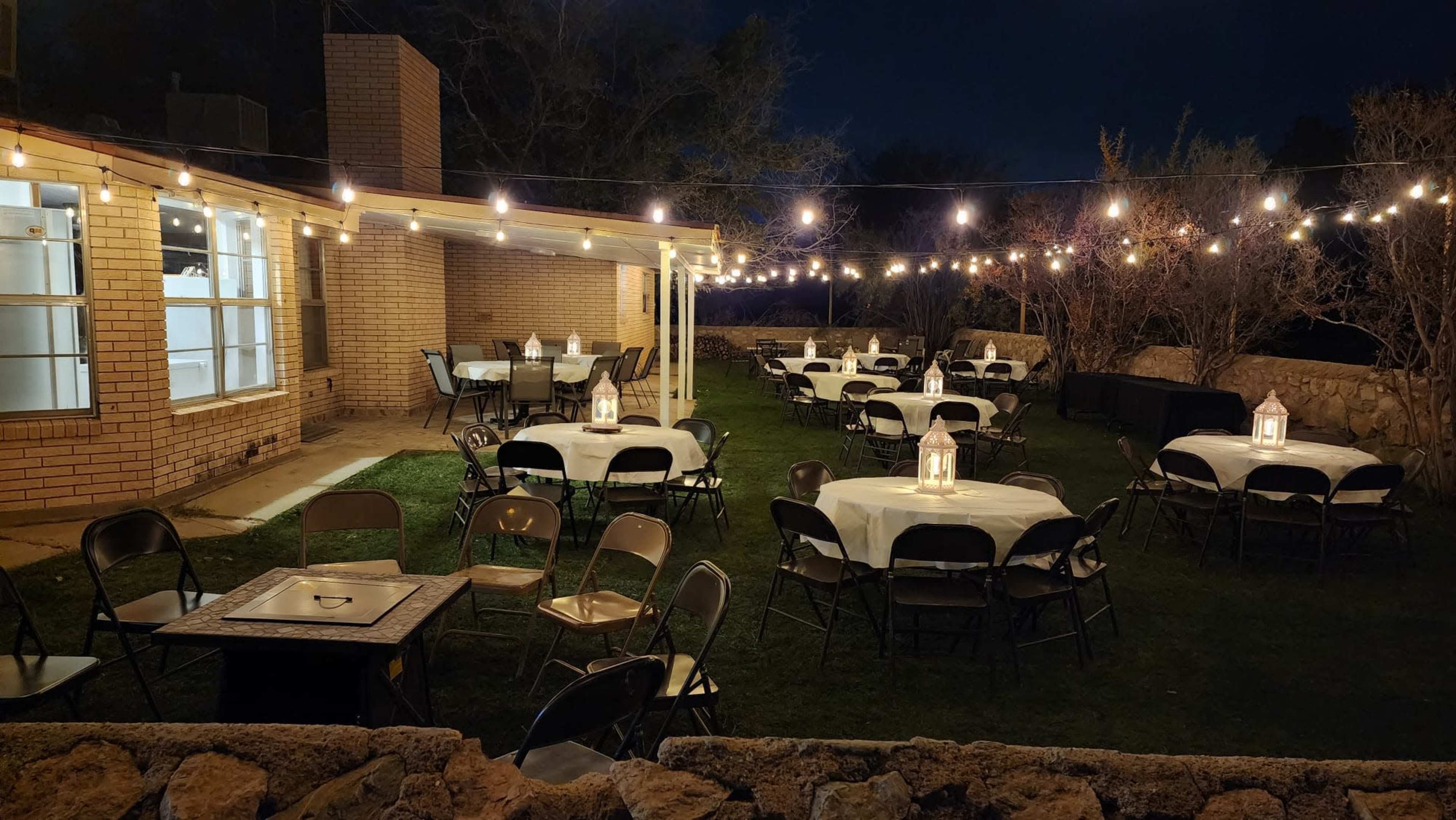 The outdoor space features several round tables set with white tablecloths under string lights, surrounded by a grass area and a brick building at night.