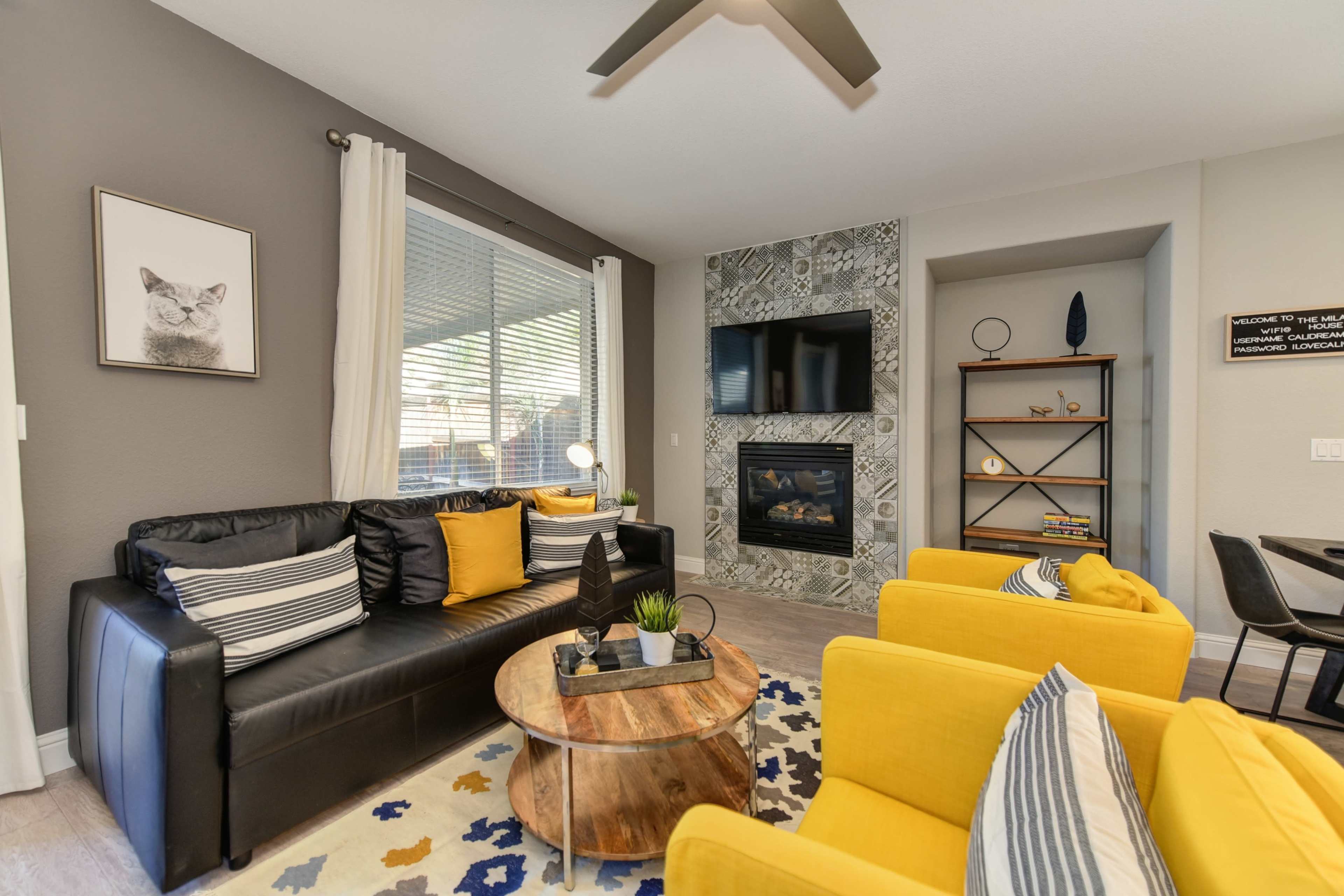 A modern living room features a black sofa with striped cushions, two yellow chairs, a round coffee table, and a fireplace surrounded by patterned tile.