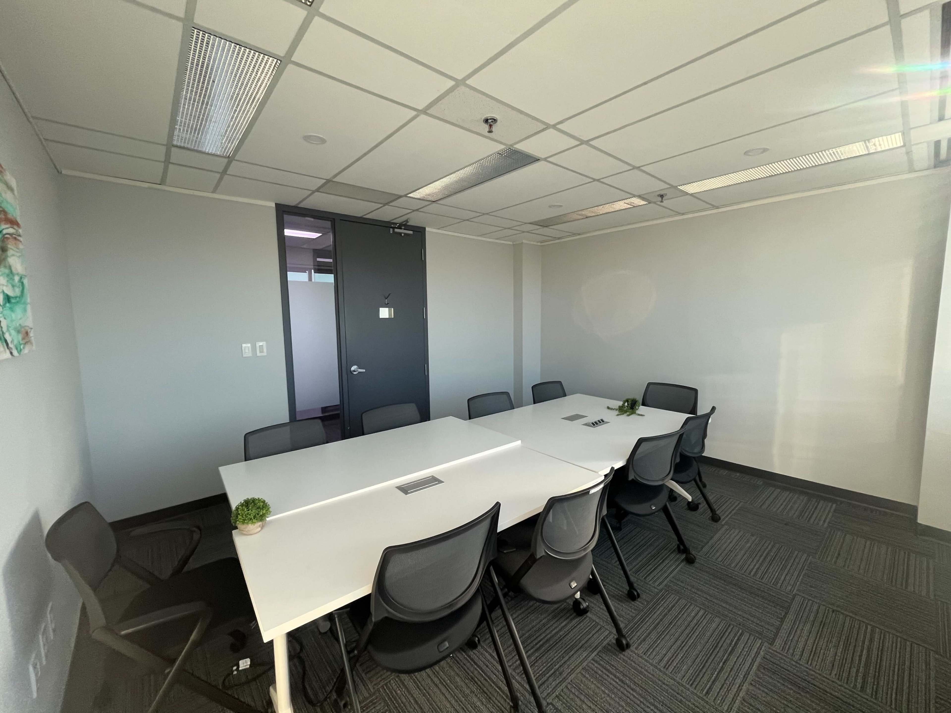 The image shows a modern conference room with a large white table surrounded by several black chairs and a large window letting in natural light.