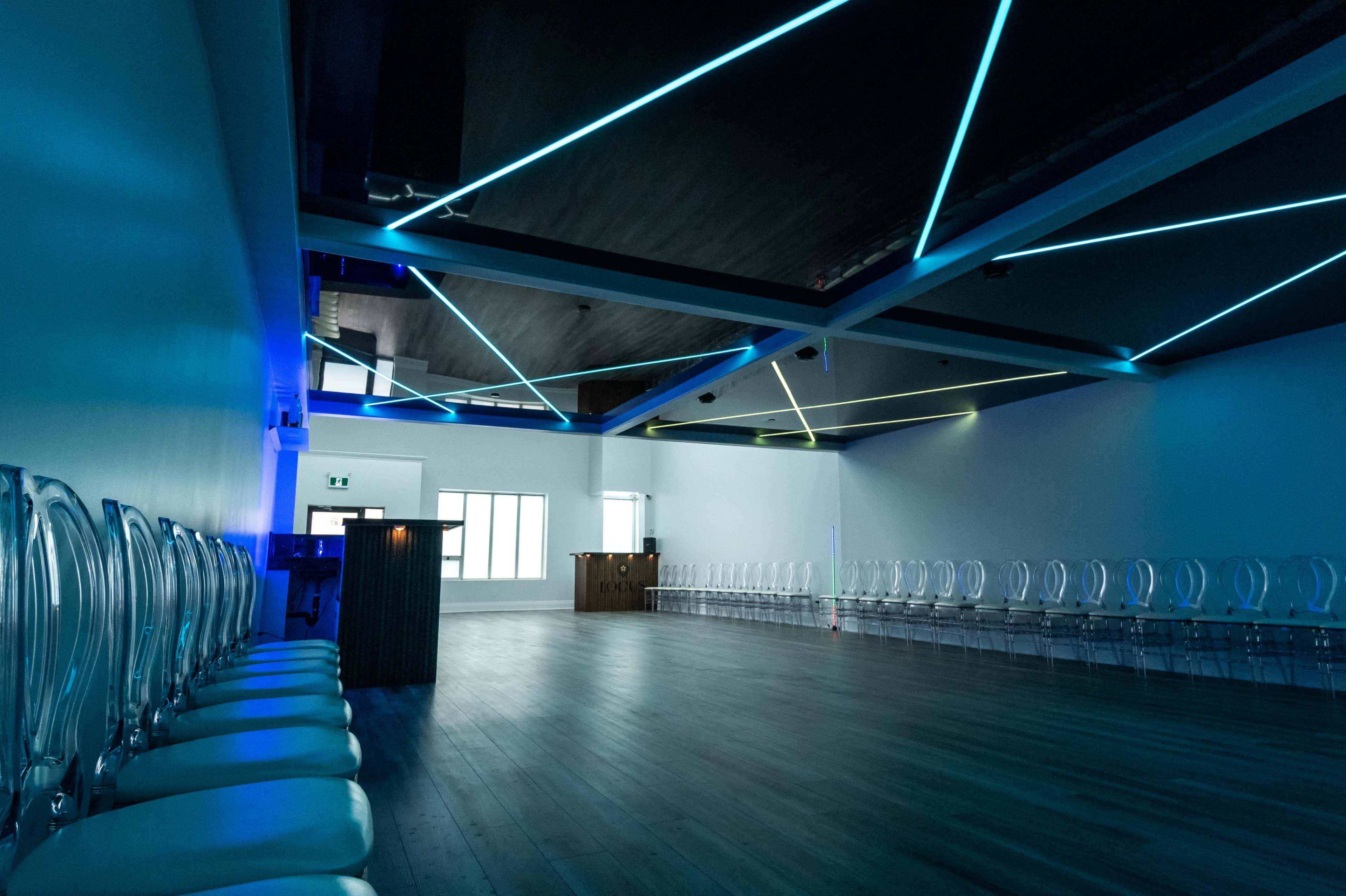 A spacious, modern room with transparent chairs arranged in rows and vivid blue and green lighting on the ceiling.