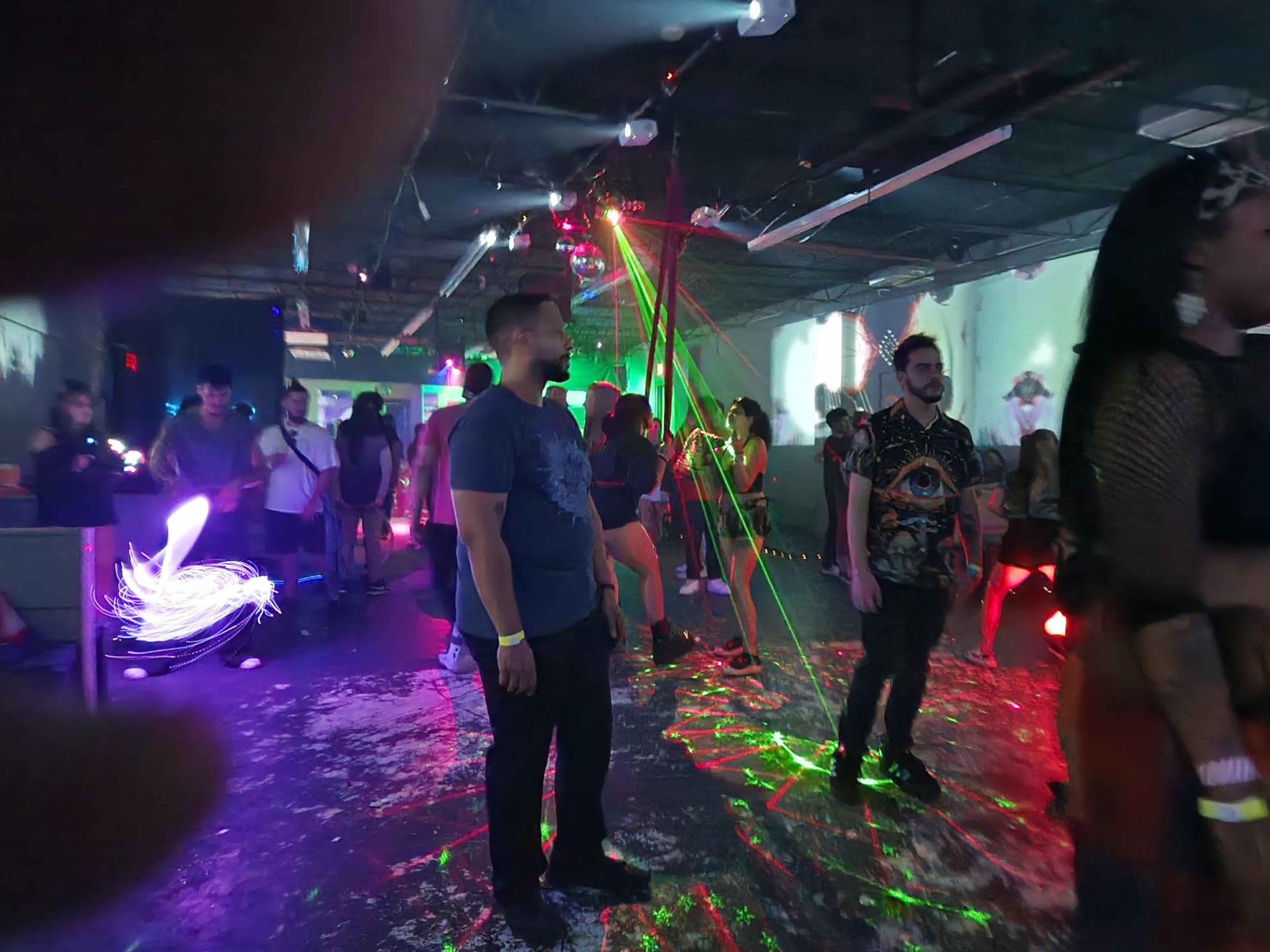 A crowd of people stands in a dimly lit space with colorful laser lights and atmospheric effects during a party or event.