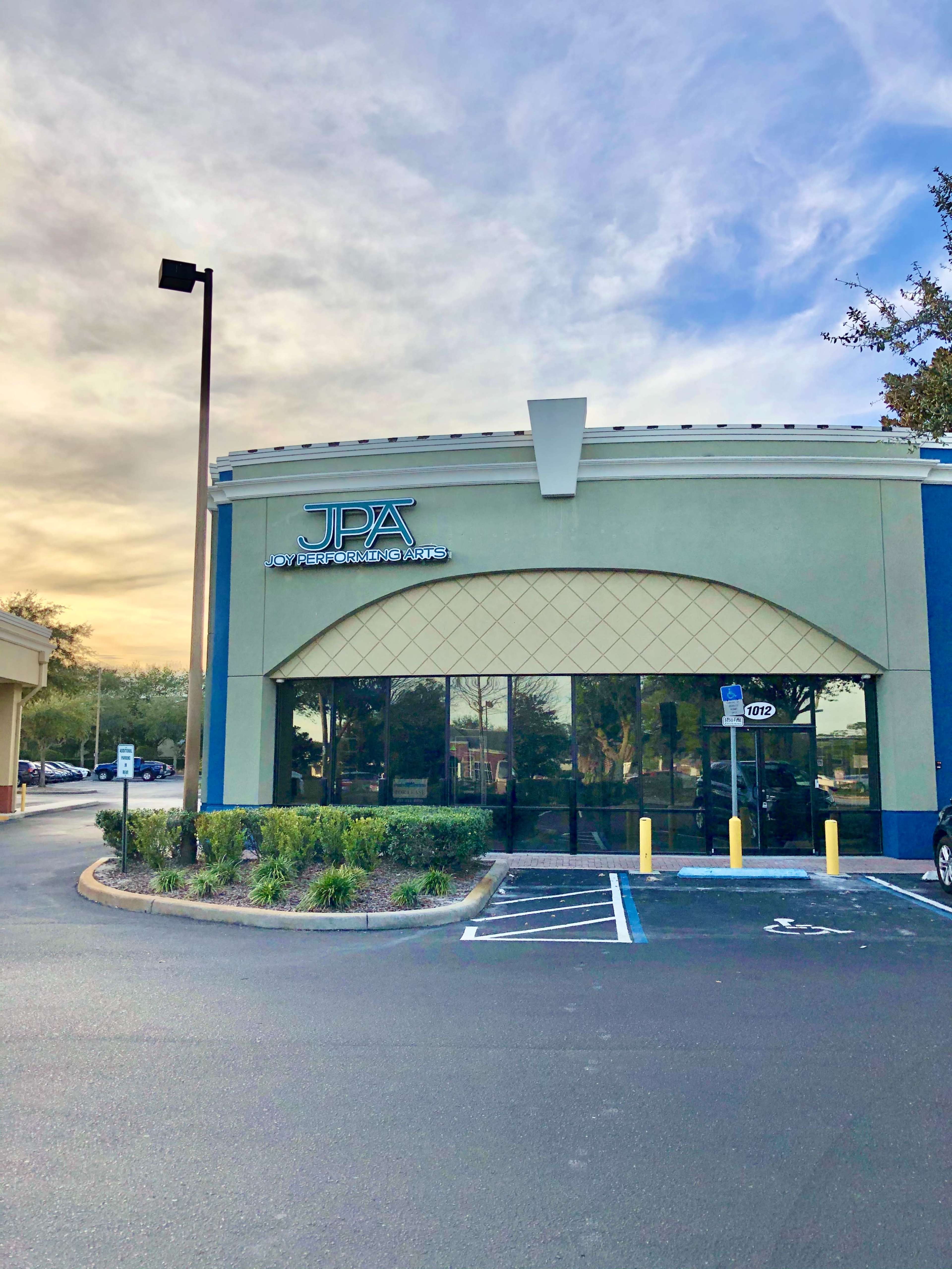 The exterior of a commercial building with the sign "JPA" prominently displayed, surrounded by landscaped areas and a parking lot under a cloudy sky.
