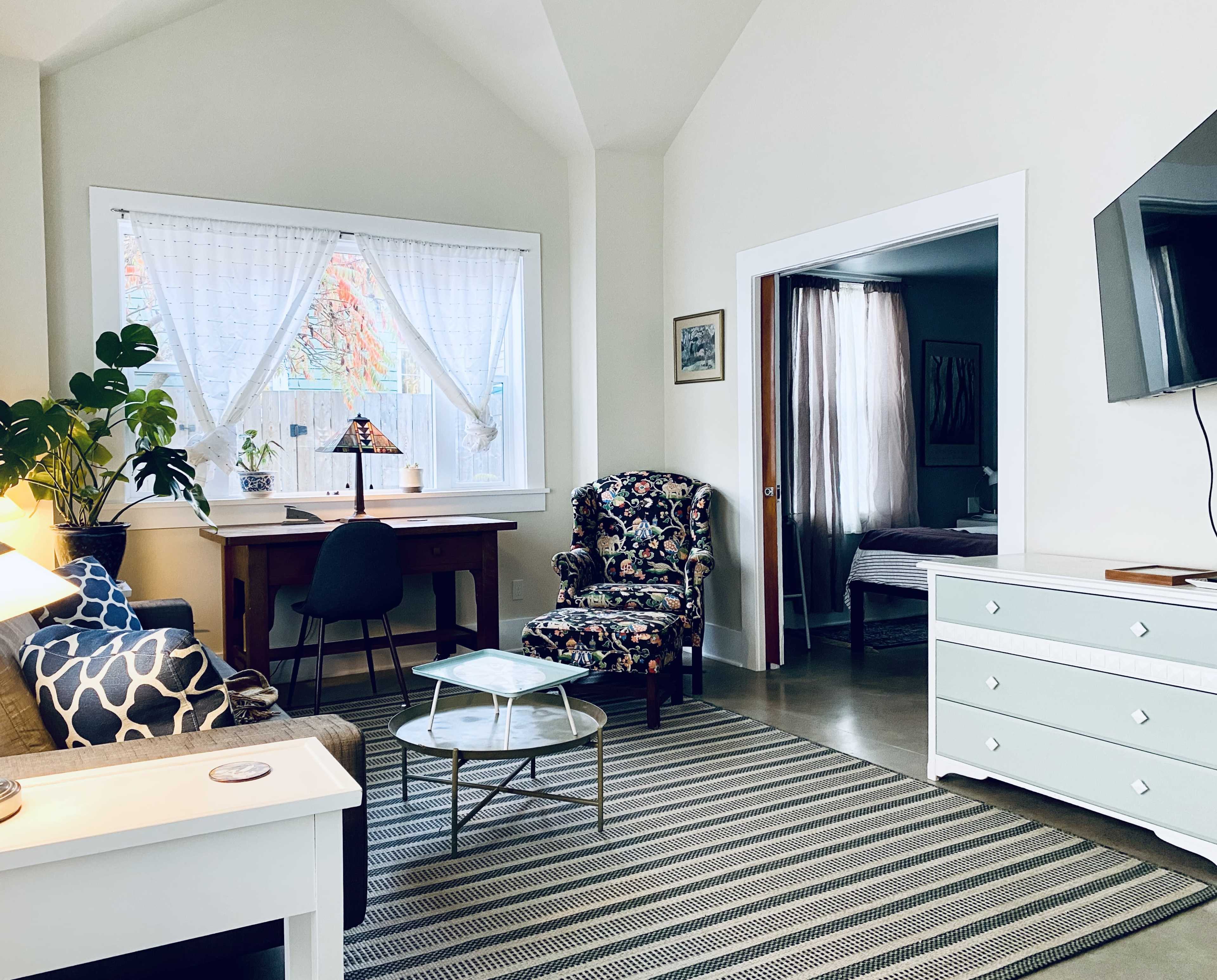 The image shows a bright living space featuring a floral armchair, a wooden desk by a window, a light-colored dresser, and a television mounted on the wall.