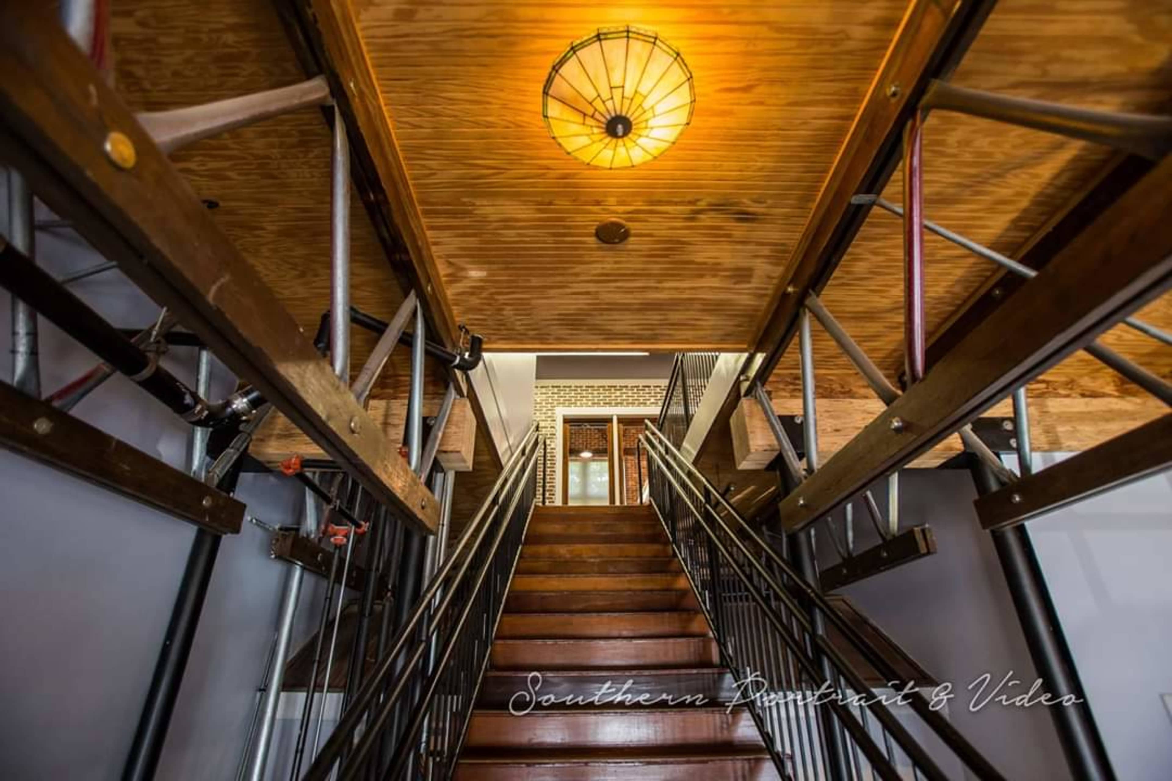 A wooden staircase leading up to a landing, illuminated by a ceiling light, with metal railing and exposed beams in the surroundings.