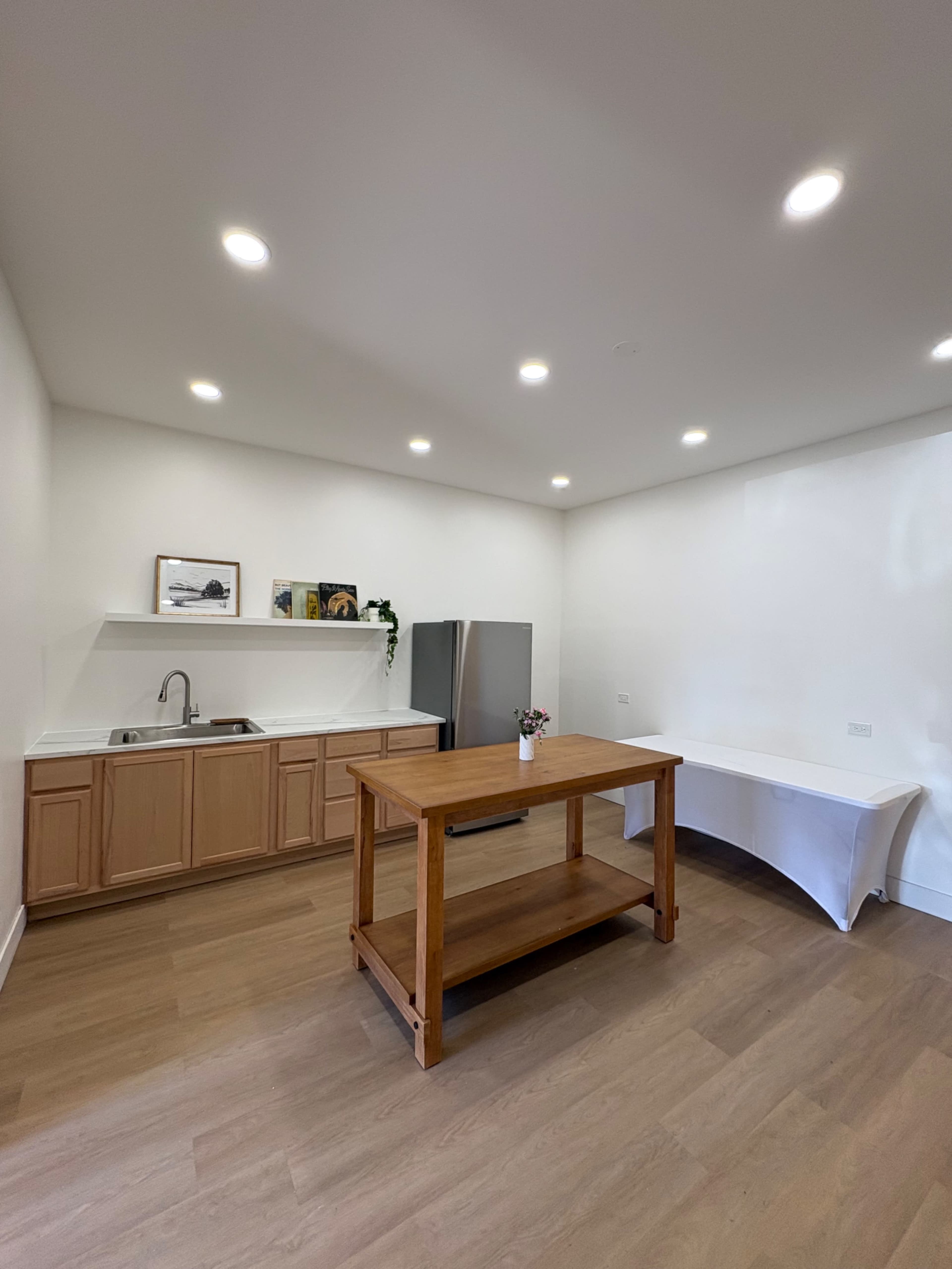 The image shows a minimalist kitchen with a wooden table in the center, a sink and countertop on one side, and a refrigerator against the wall.