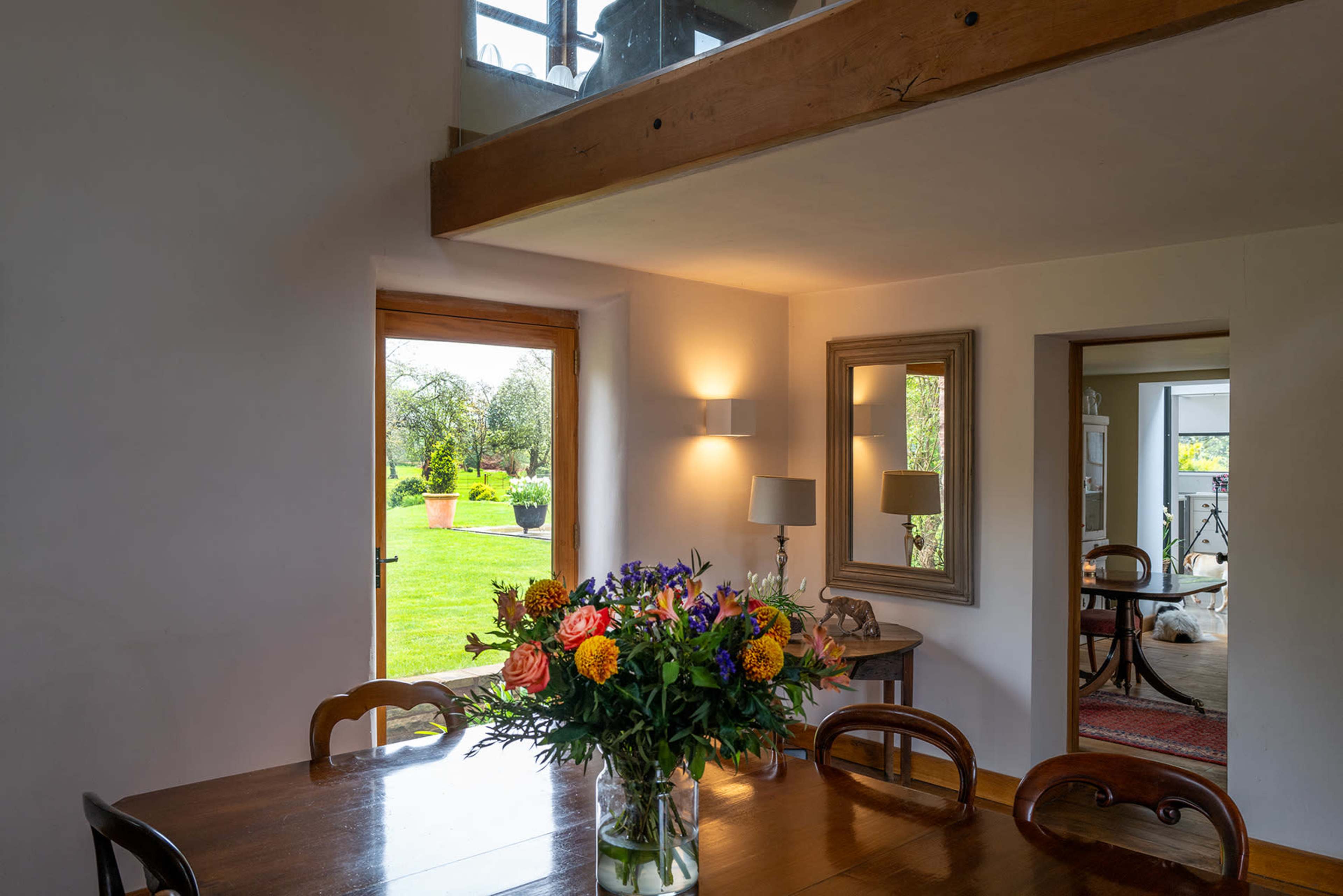 A dining area features a wooden table with a floral arrangement, adjacent to a door leading to a garden and a doorway to another room.