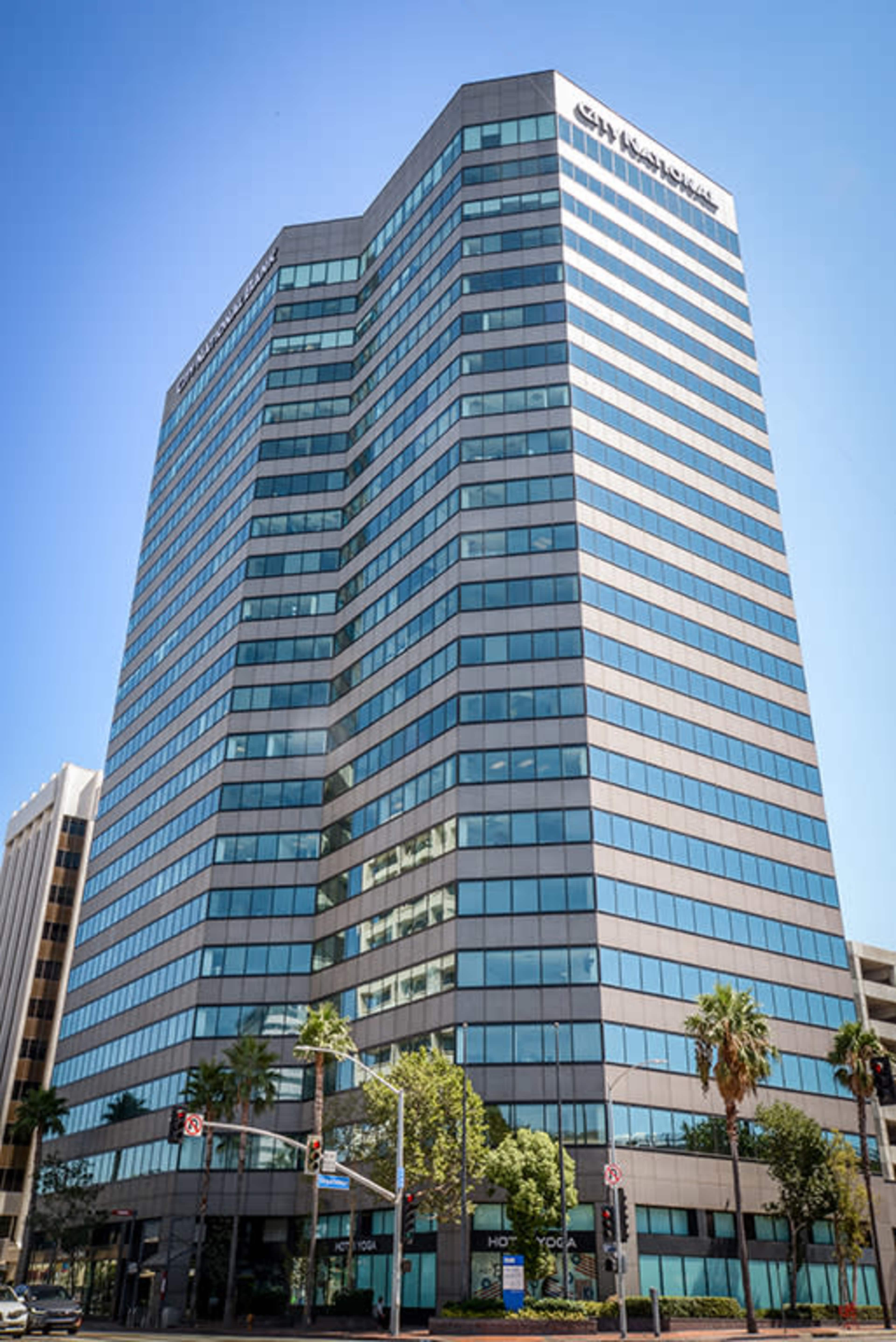 A tall glass office building is seen on a sunny day, surrounded by palm trees and city streets.