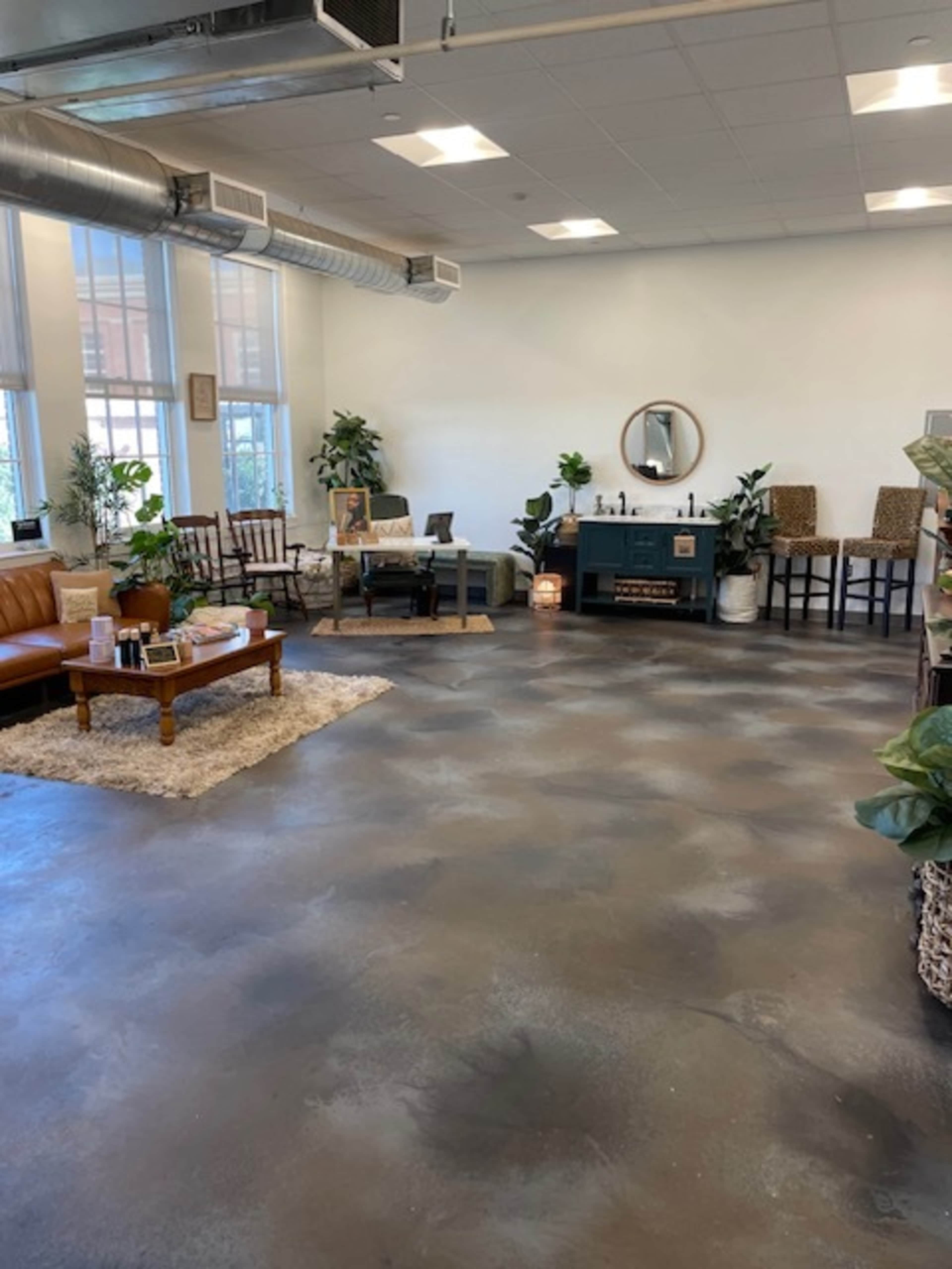A spacious room with a concrete floor, furnished with a brown leather couch, wooden chairs, a coffee table, and a dark cabinet, surrounded by indoor plants.