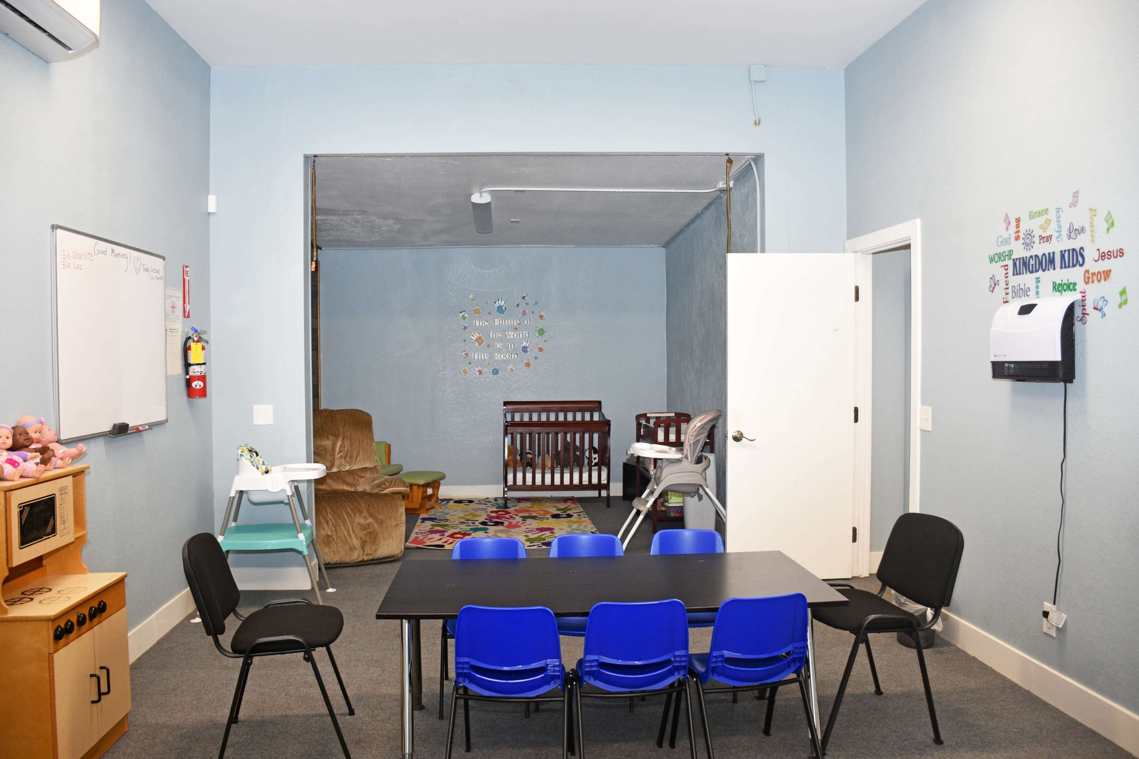 A brightly lit classroom with a black table surrounded by blue chairs, a doorway leading to a cozy seating area, and a nursery setup with a crib and brightly colored rug.
