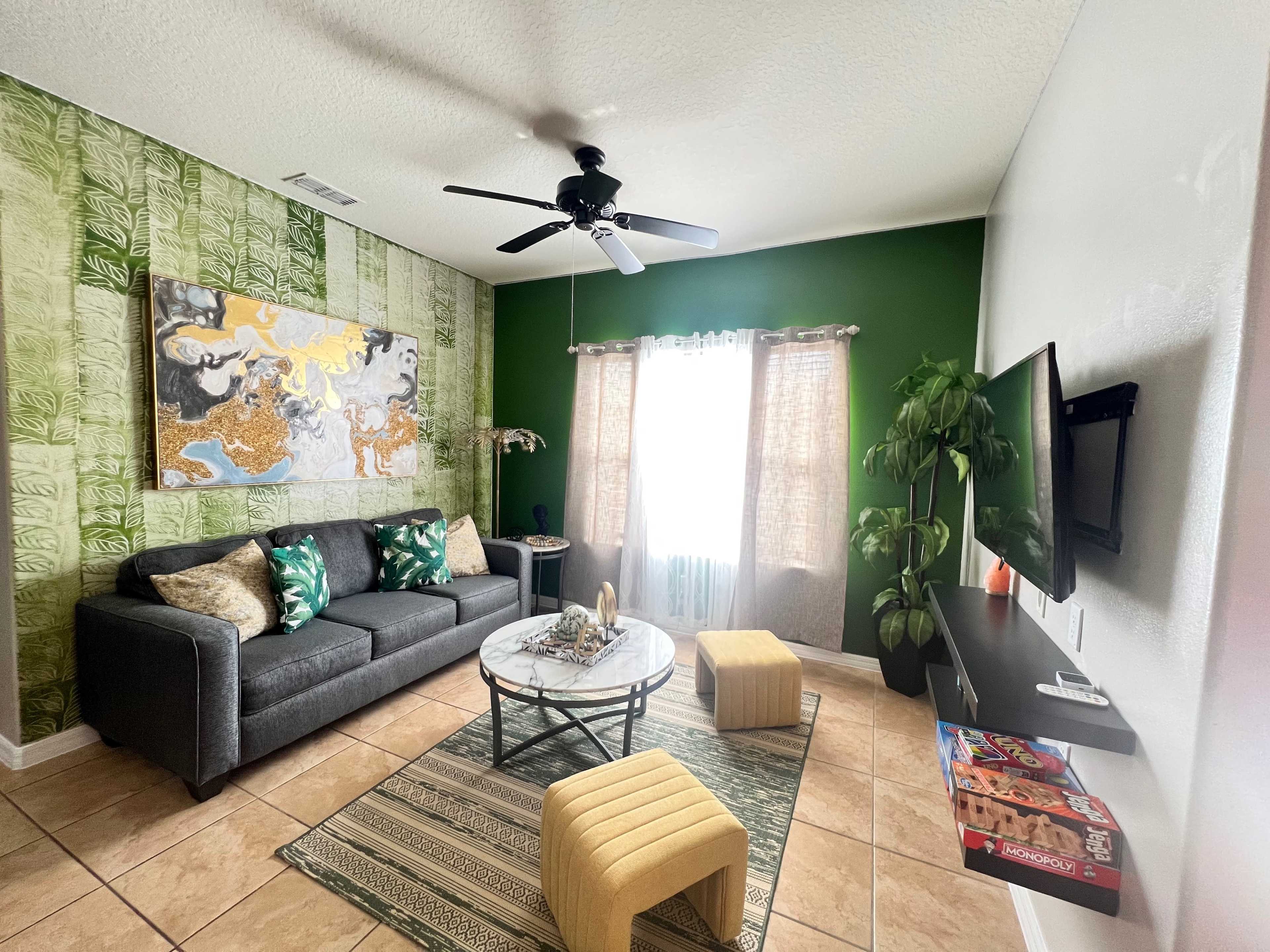 The living room features a gray sofa, a round coffee table, a large colorful wall painting, and a green accent wall with natural light coming through the sheer curtains.