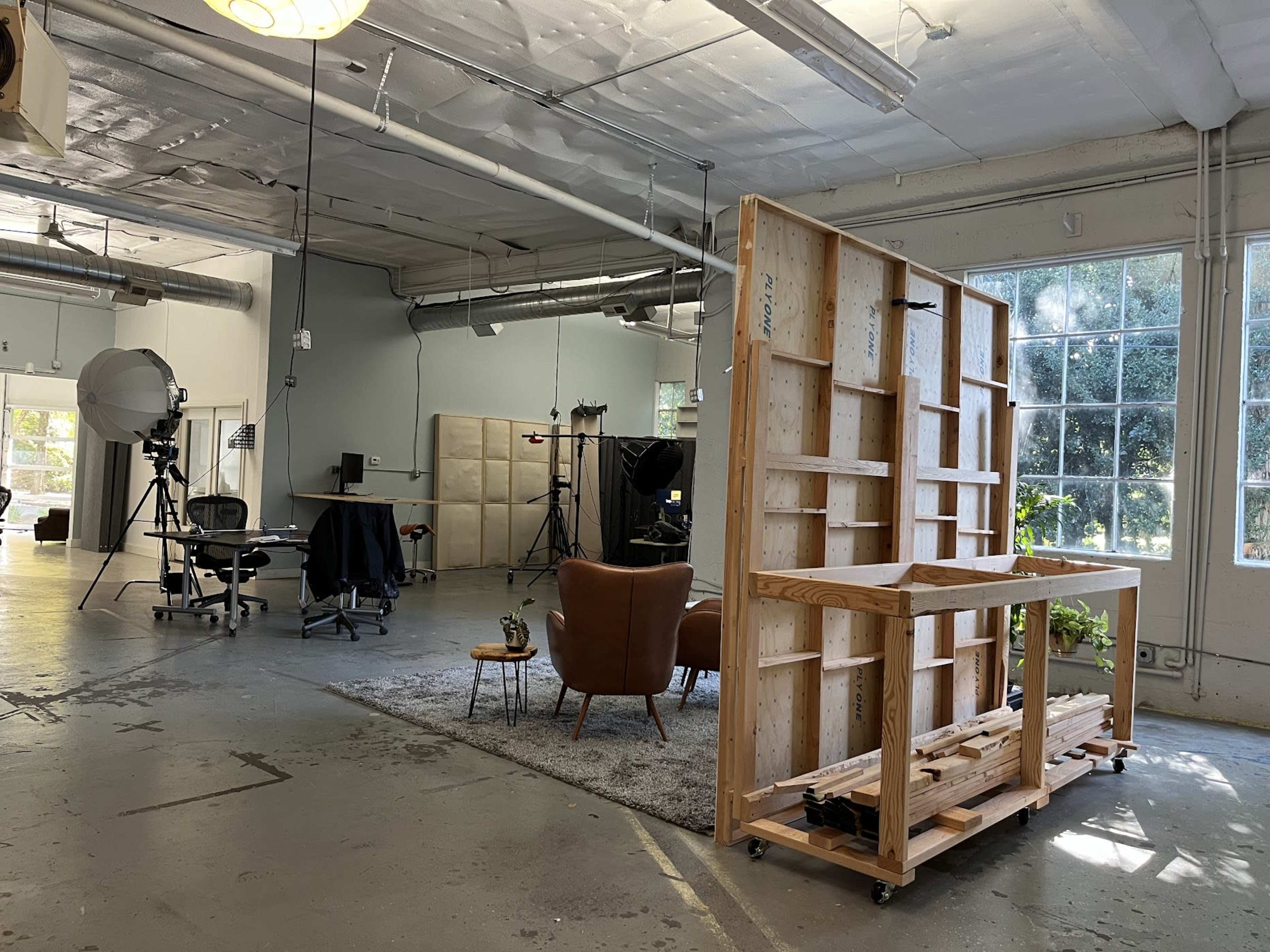 A spacious studio with a seating area, large windows, a wooden shelving unit, and various filming equipment set up in the background.