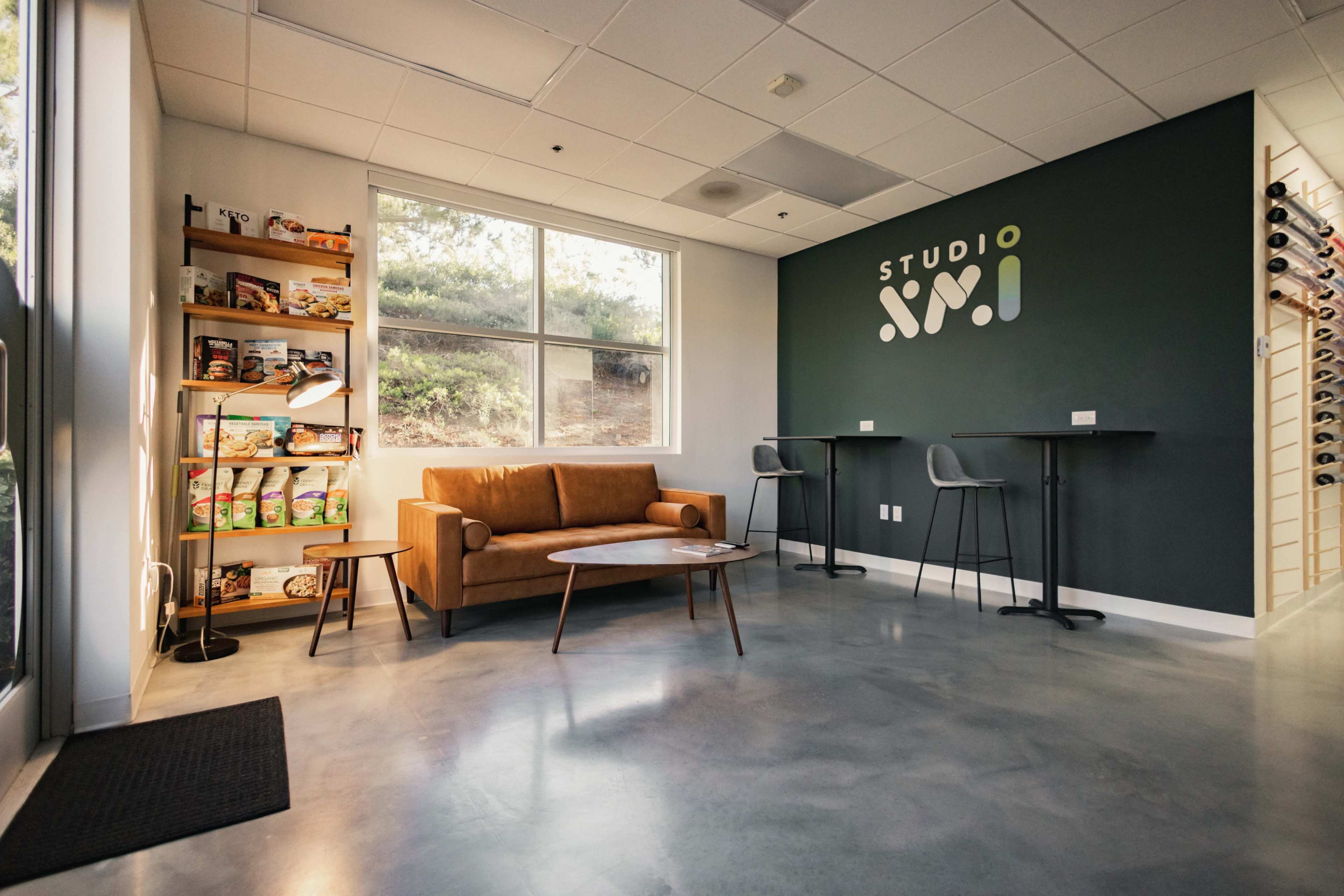 A modern room with a brown couch, a minimalist table, and a wall featuring the text "STUDIO XM," along with a shelf filled with snacks and drinks.