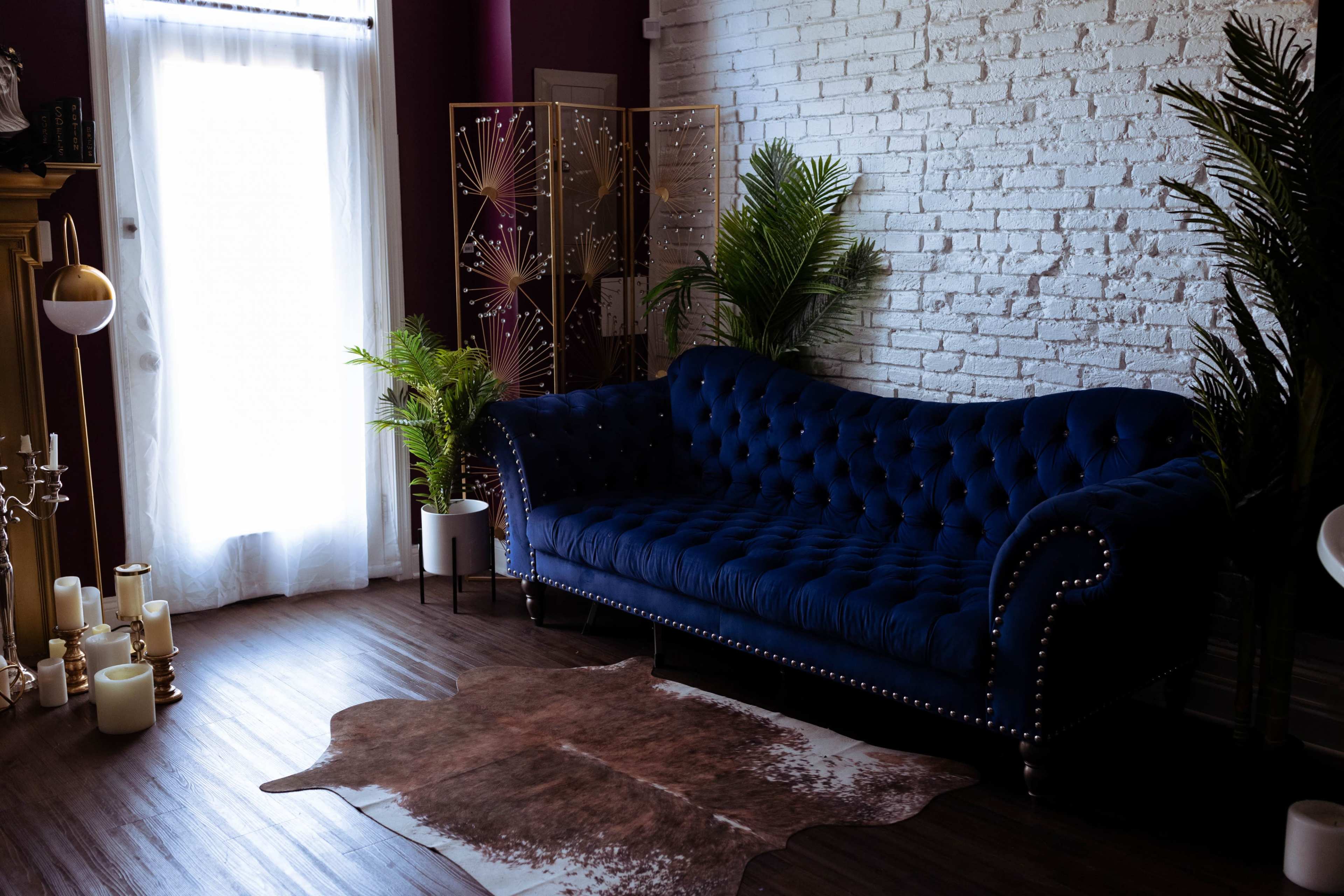 The room features a blue tufted sofa, a brown cowhide rug, potted plants, and a decorative folding screen against a white brick wall.