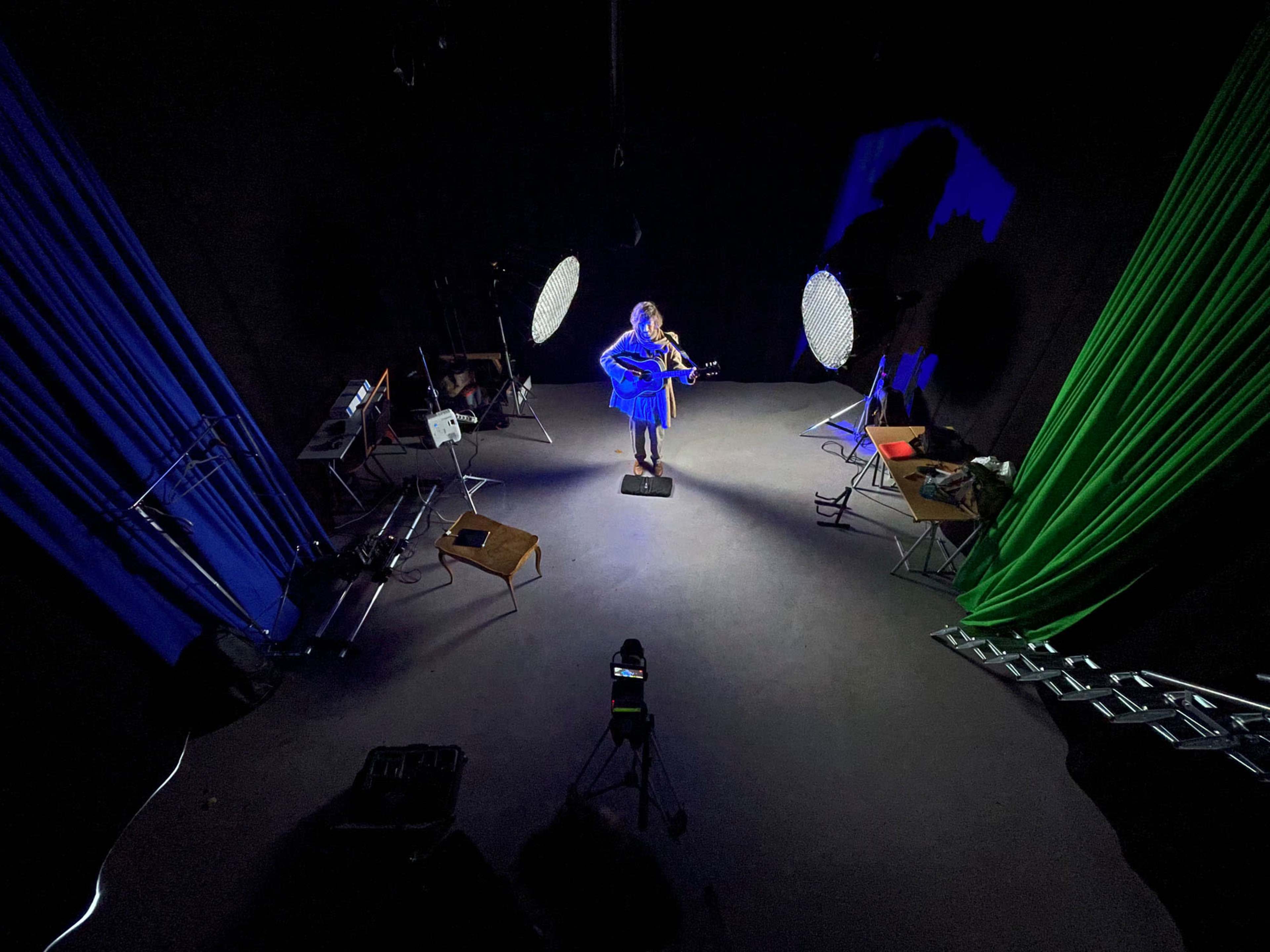 A musician stands on a stage with a guitar in a dimly lit studio surrounded by colorful backdrop curtains and lighting equipment.