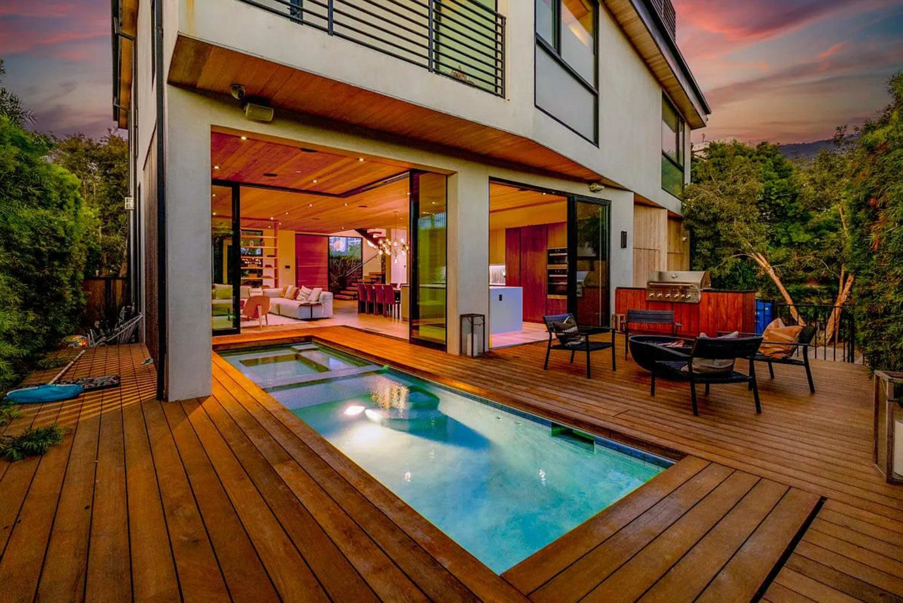The image shows a modern house with large glass doors opening to a wooden deck and a swimming pool, surrounded by greenery and a colorful sunset.