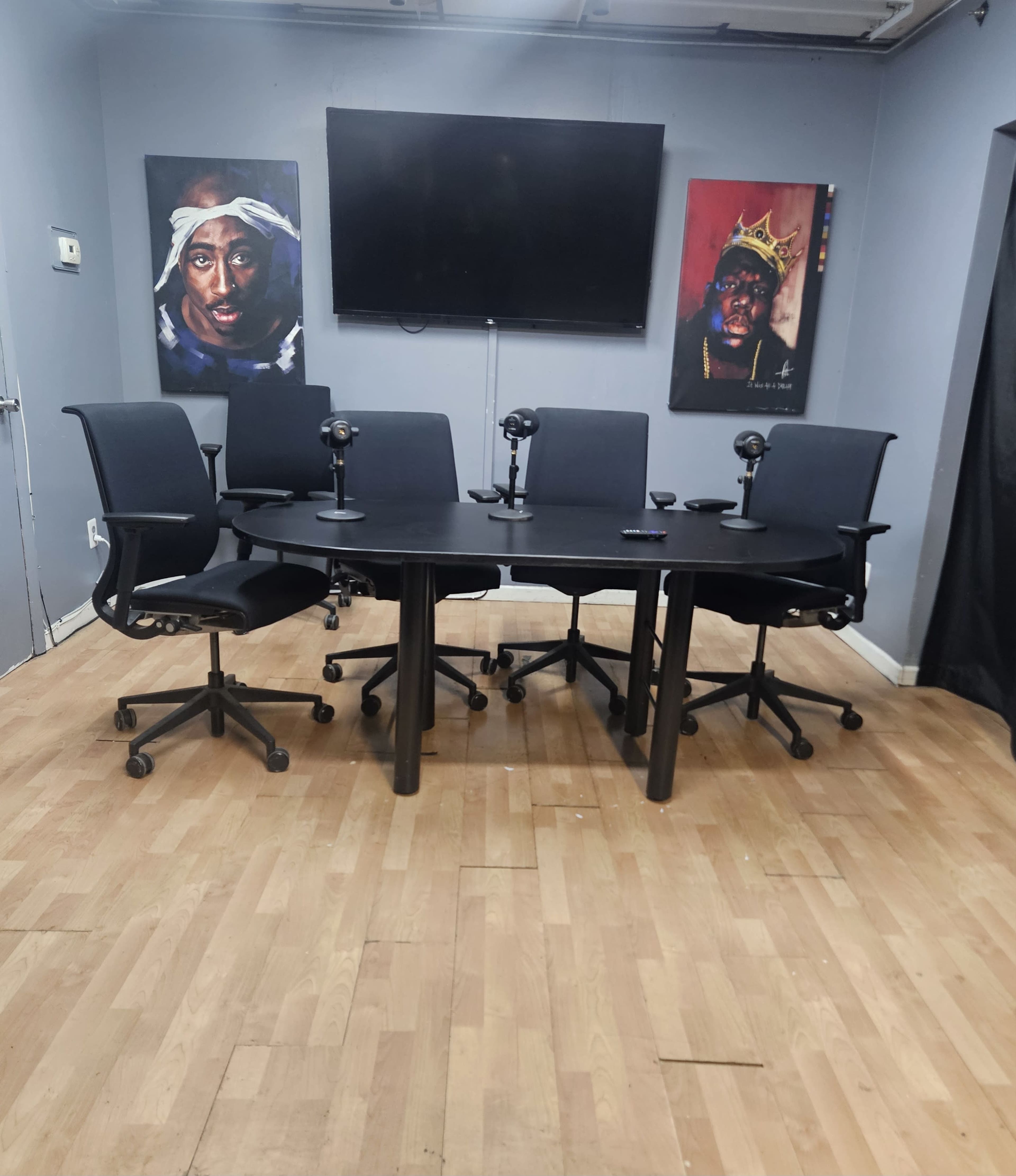 A round table with five black chairs and microphones is positioned in a room featuring two portraits on the walls and a mounted television.