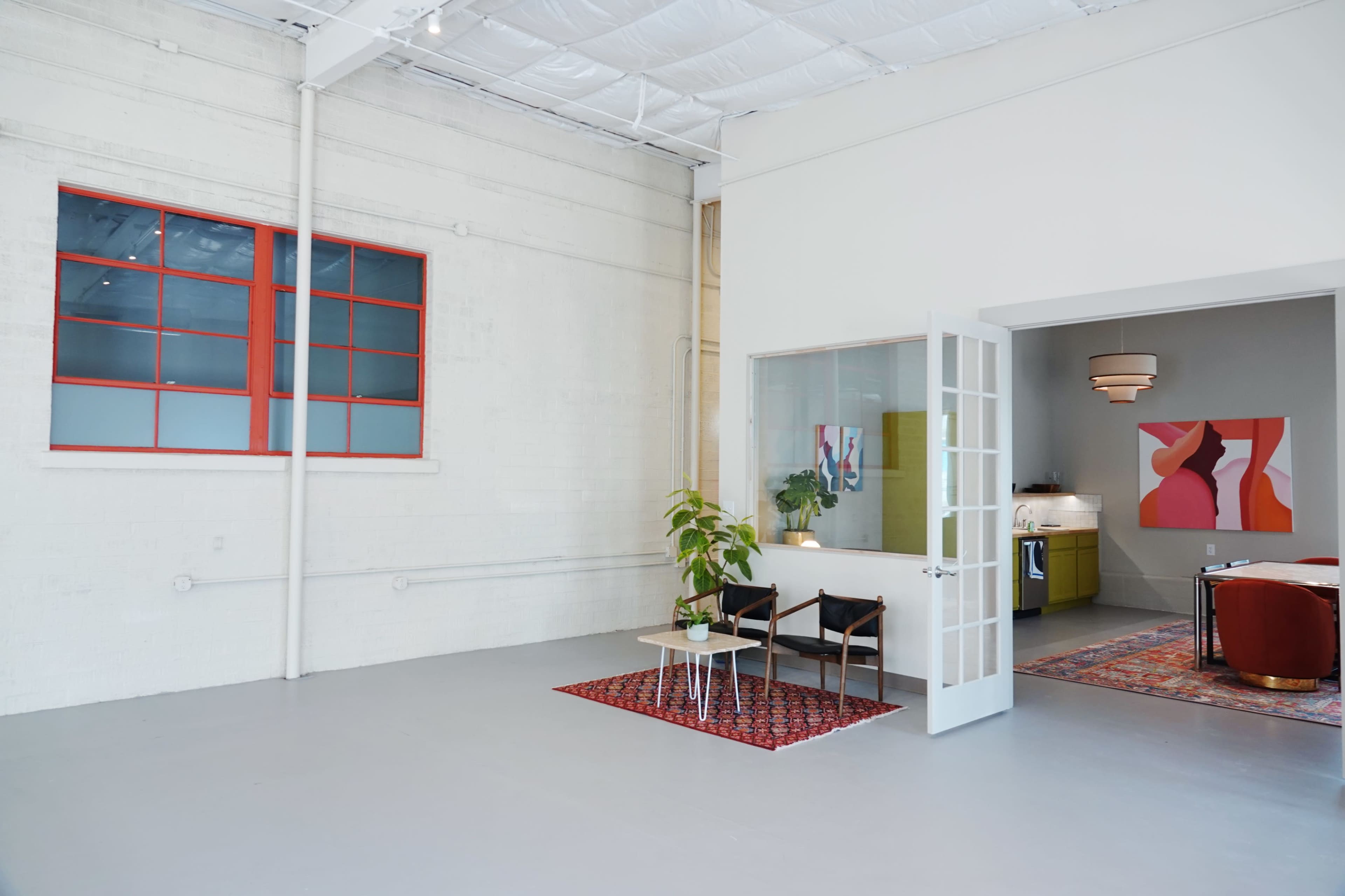 A spacious, modern interior with white walls, large windows, and a small seating area featuring a plant and a rug, leading to a separate section that includes a kitchenette.