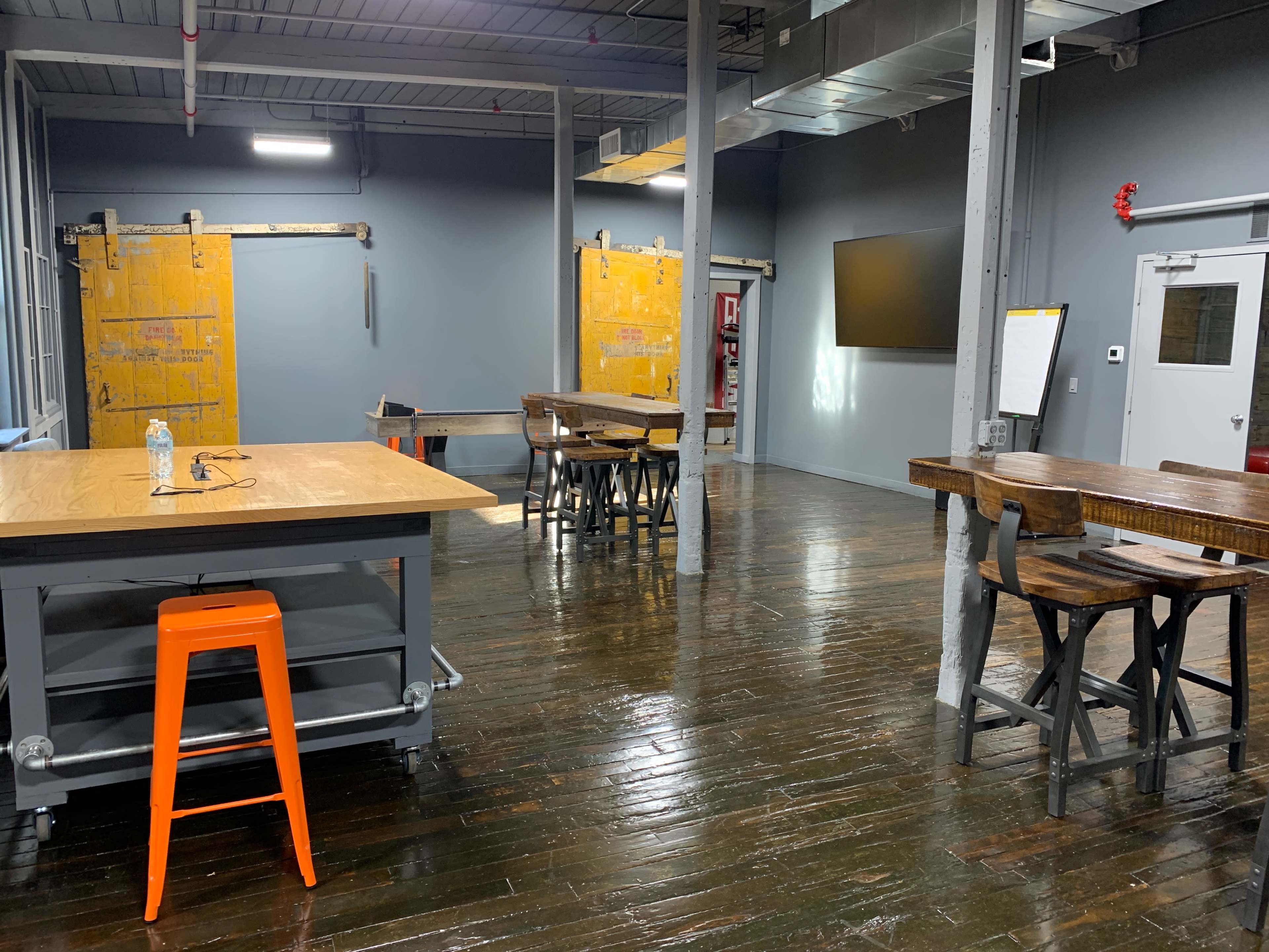 The image shows a spacious, industrial-style room with wooden tables and a large television mounted on the wall.
