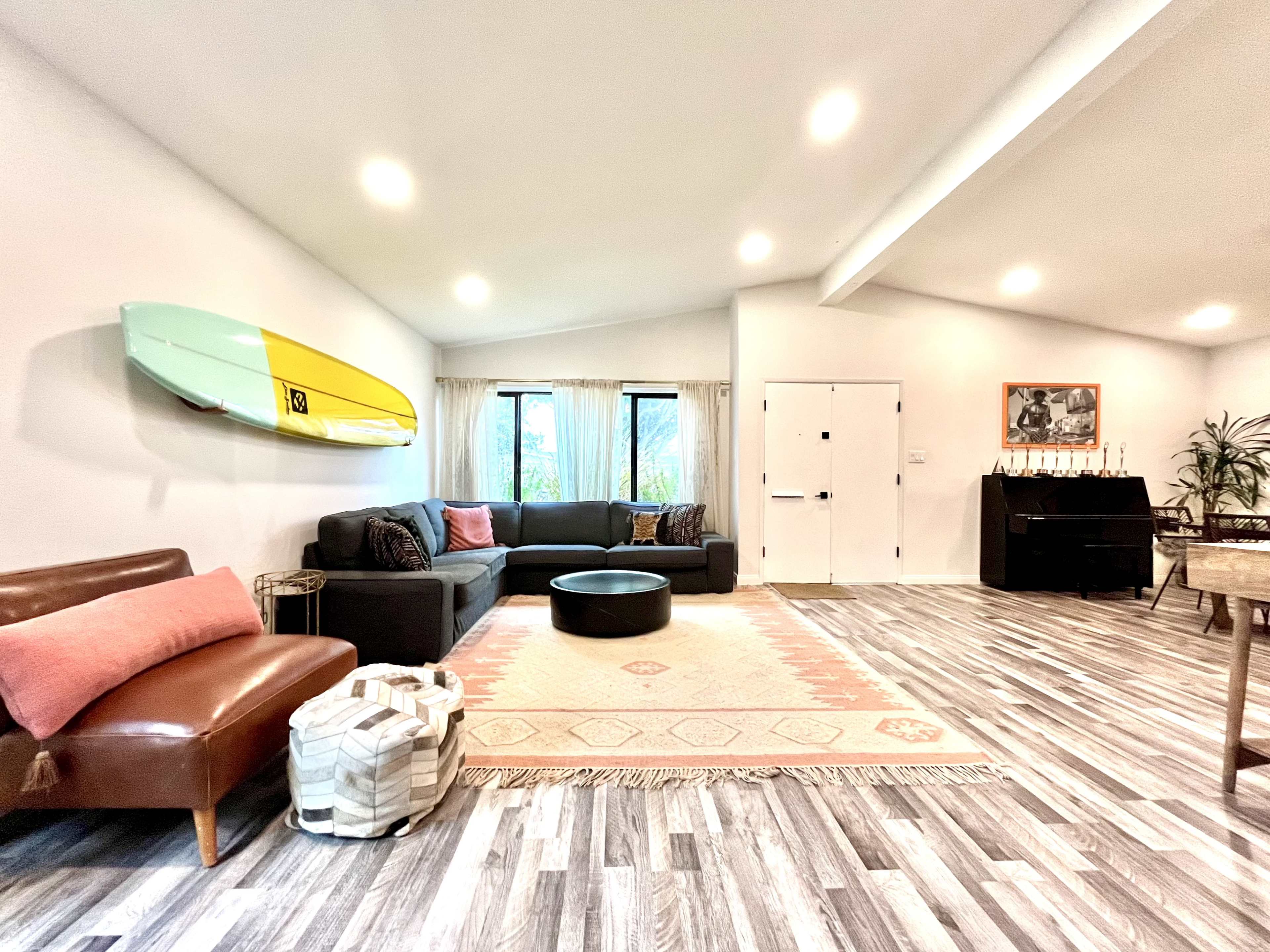 A spacious living room features a sectional sofa, a round coffee table, a patterned rug, and a surfboard mounted on the wall.