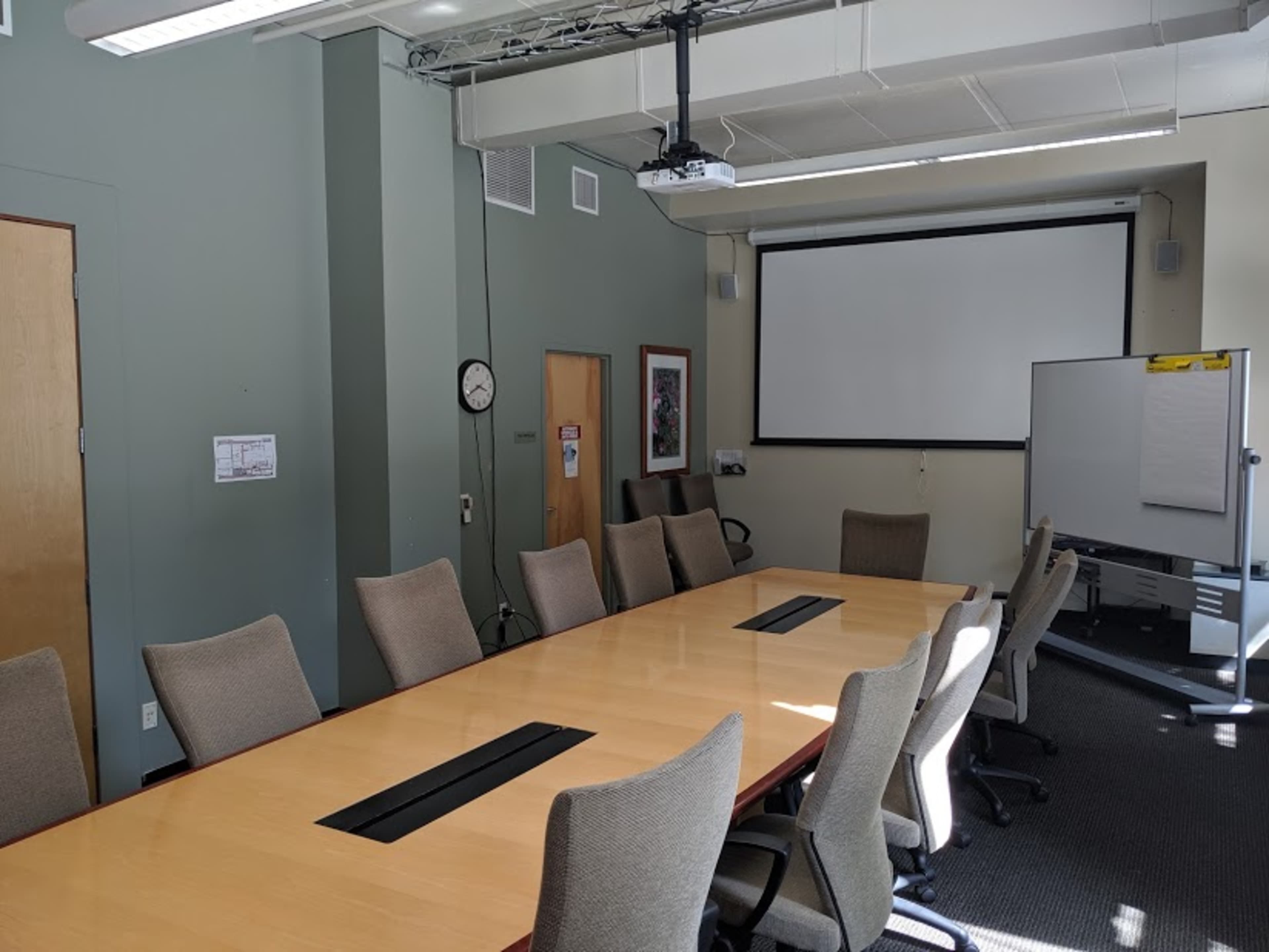 A conference room features a long table surrounded by chairs, a projector screen on the wall, and a whiteboard in the corner.