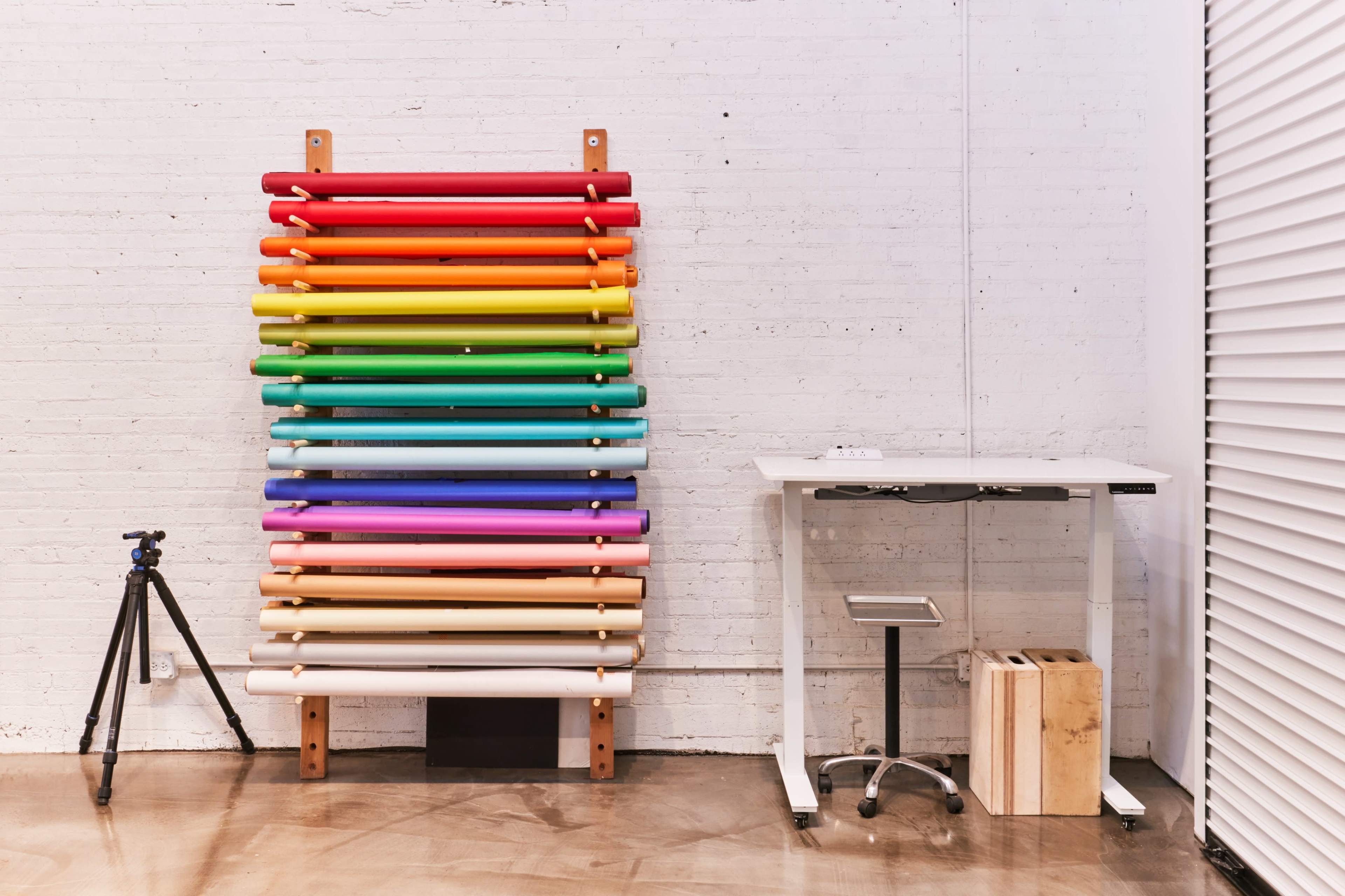 A vertical rack displays rolls of colored paper next to a white standing desk and a tripod on a concrete floor.
