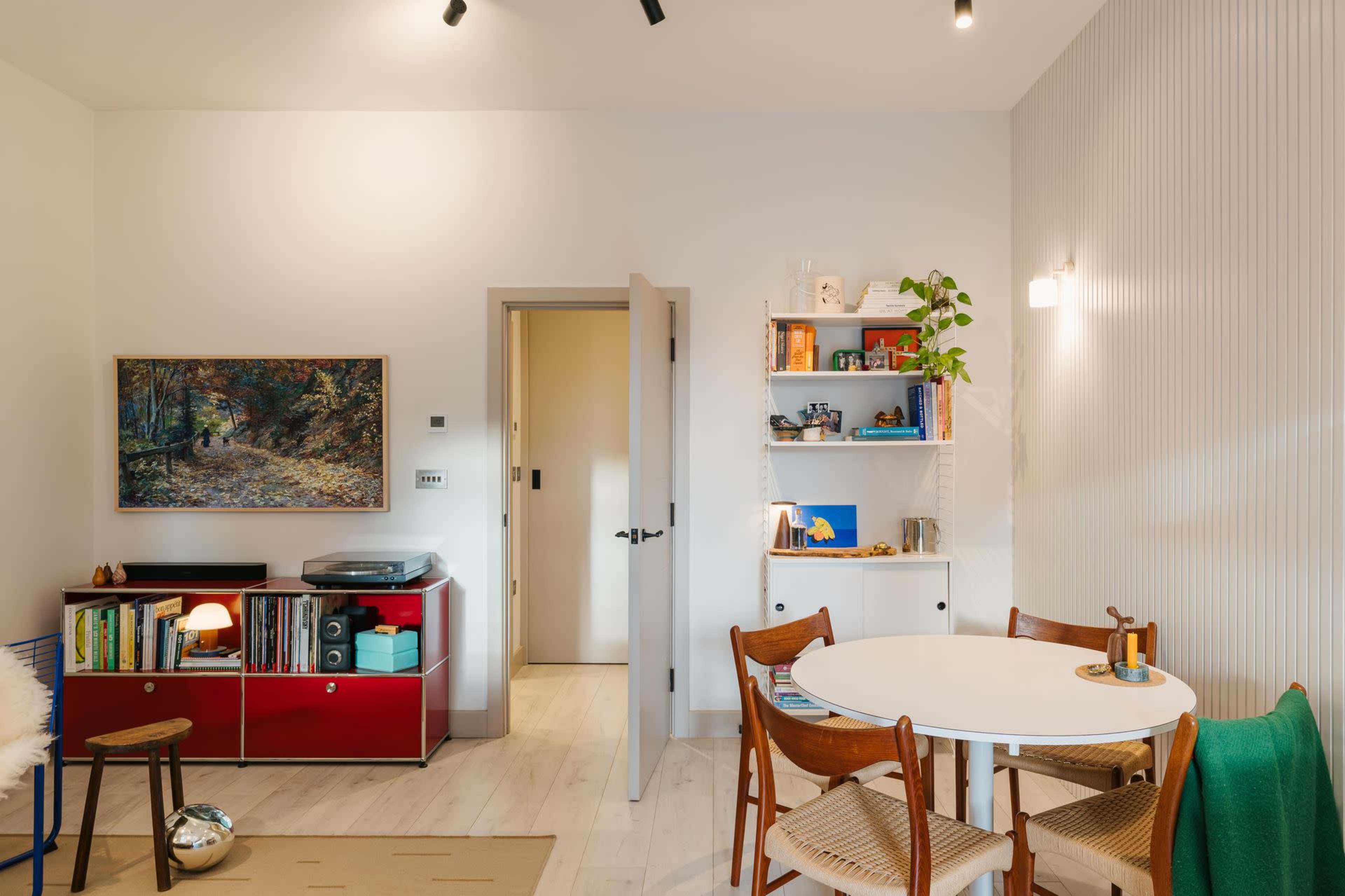 The image shows a brightly lit room featuring a round dining table with chairs, a bookshelf, a red cabinet, and a door leading to another space.