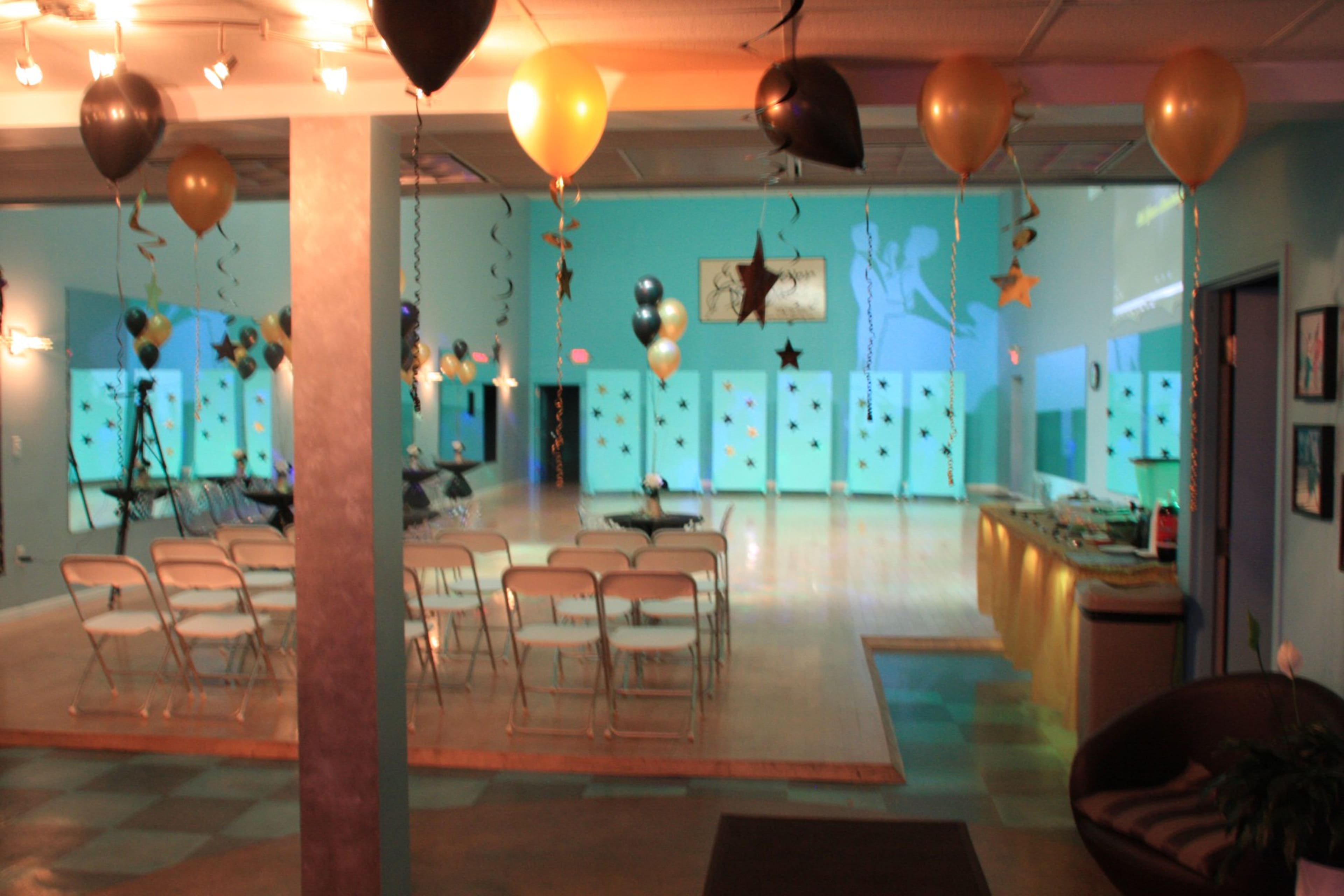 A spacious event room decorated with balloons and streamers, featuring rows of chairs set up for an audience.