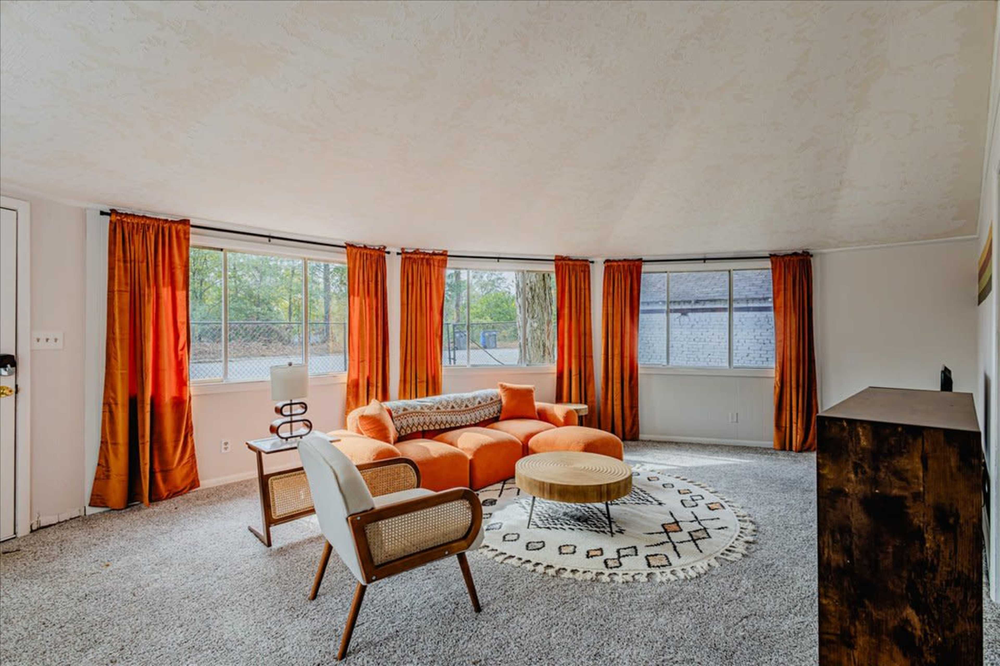 The room features a rounded design with orange curtains, a circular area rug, a modern orange sofa, and two chairs arranged around a coffee table.