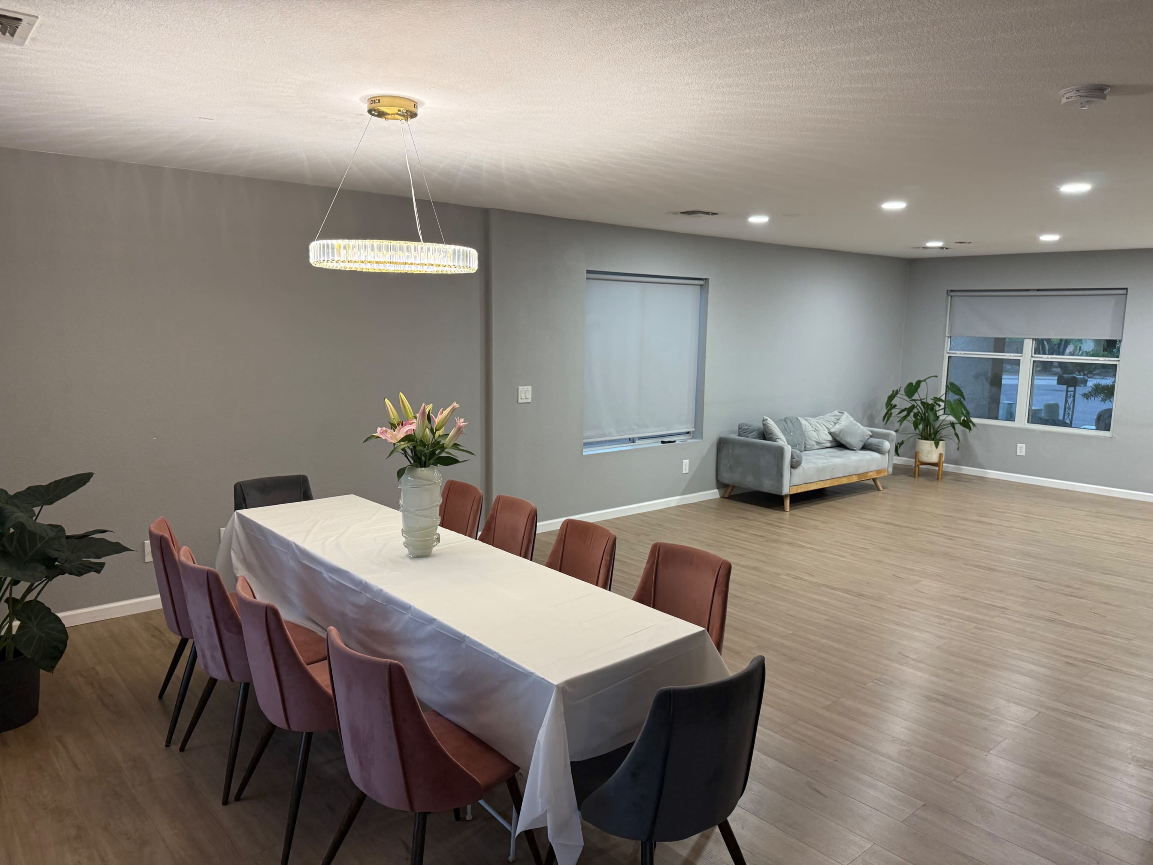 The image shows a modern dining area with a long table set for eight, a vase of flowers at the center, and a grey sofa in a well-lit living space.