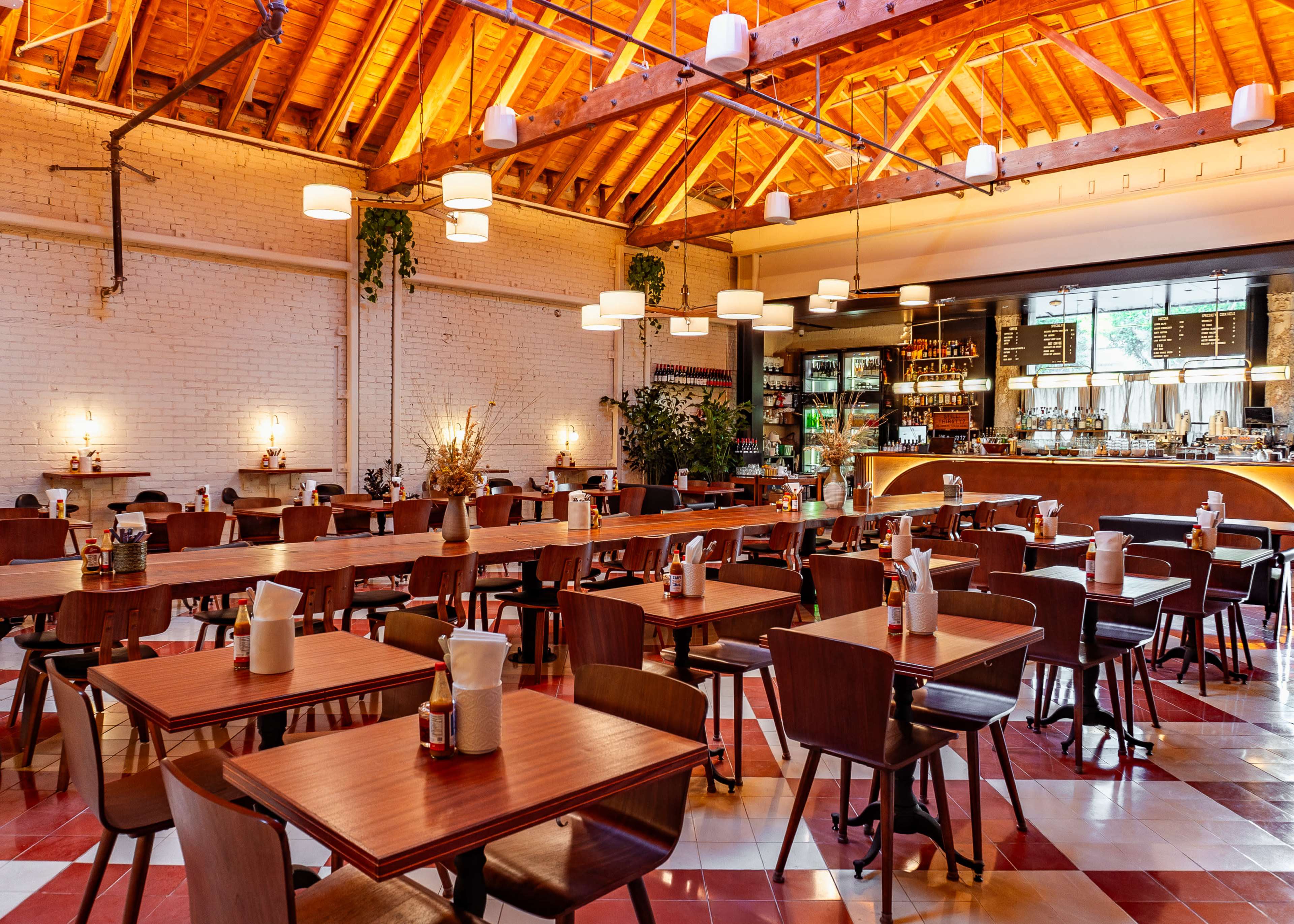 The image shows a spacious, modern restaurant with wooden tables and chairs arranged neatly, featuring a bar area with a variety of beverages in the background.